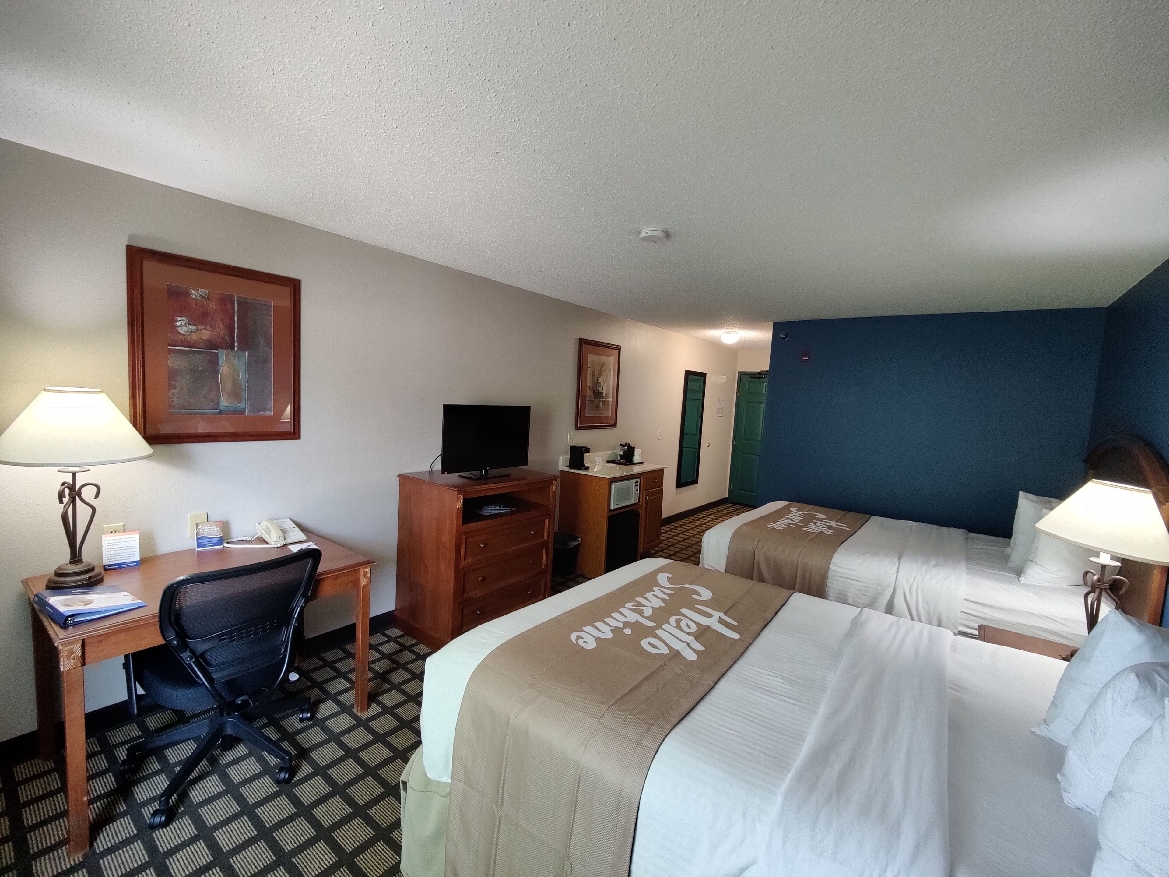 A hotel room with two beds, a desk with a computer, a chair, a TV, and a small kitchenette.