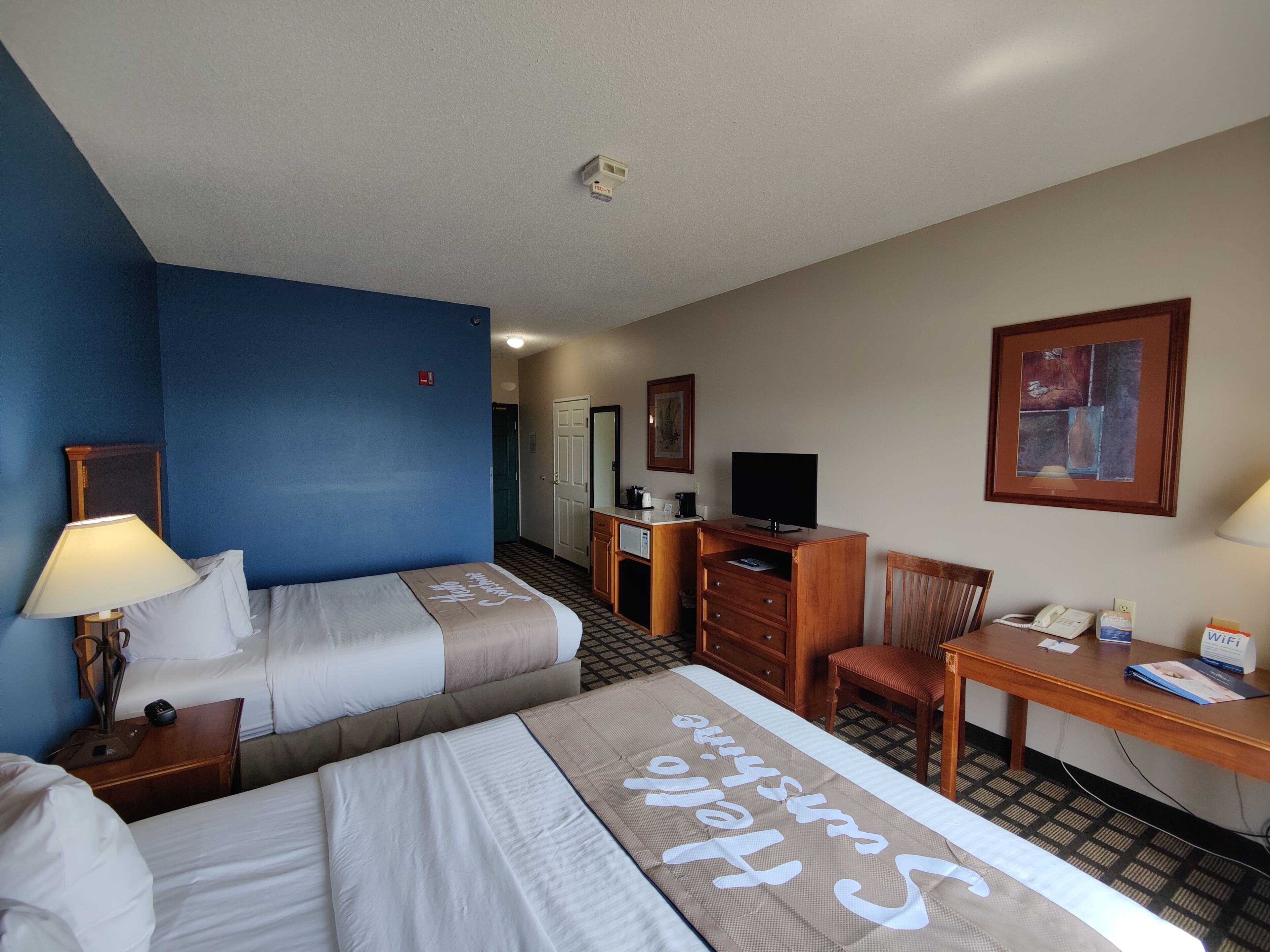 A hotel room with two beds, a desk, a chair, a television, and a painting on the wall.