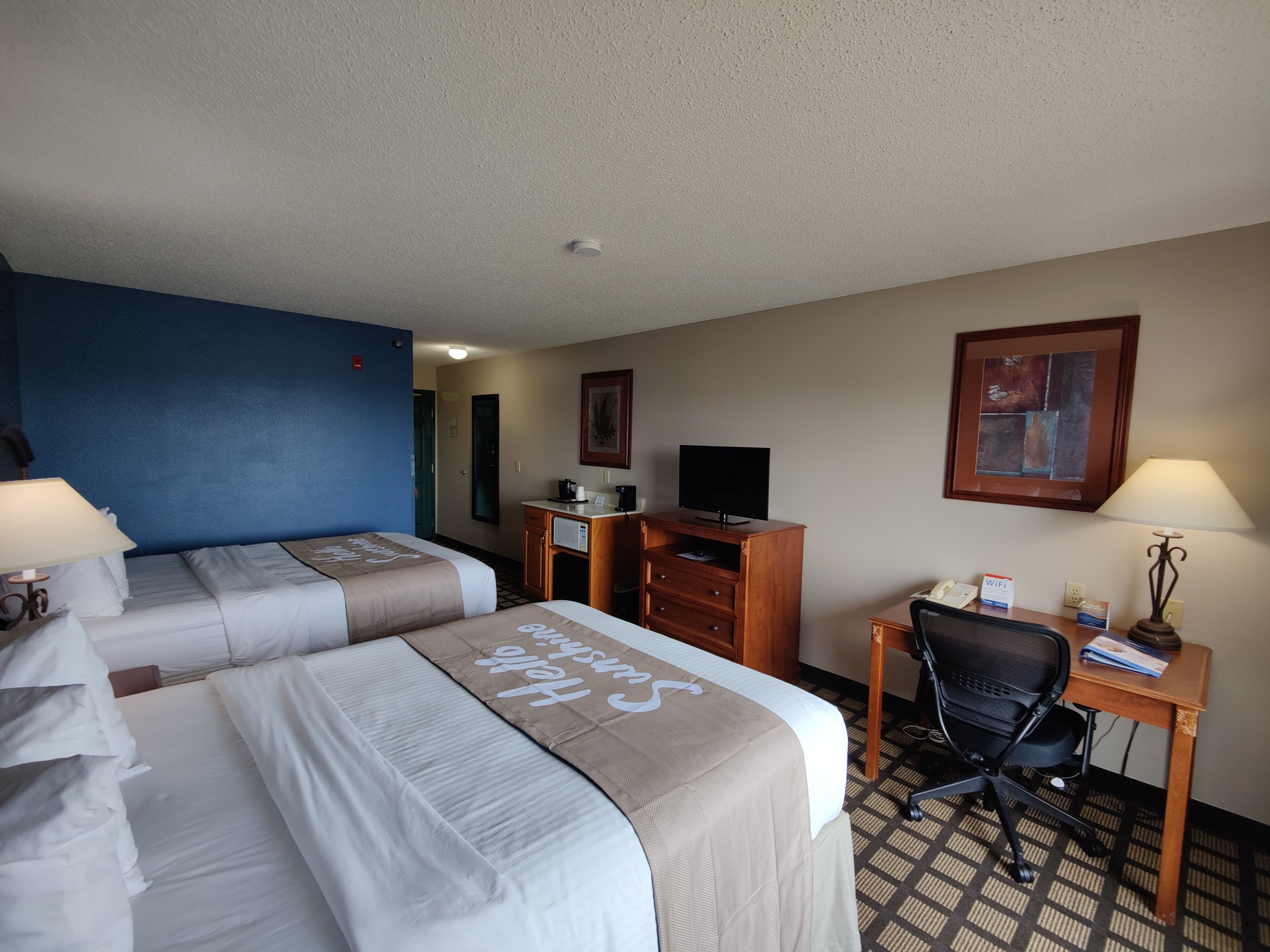 Habitación de hotel con dos camas, un escritorio, una silla, un televisor y un cuadro en la pared.
