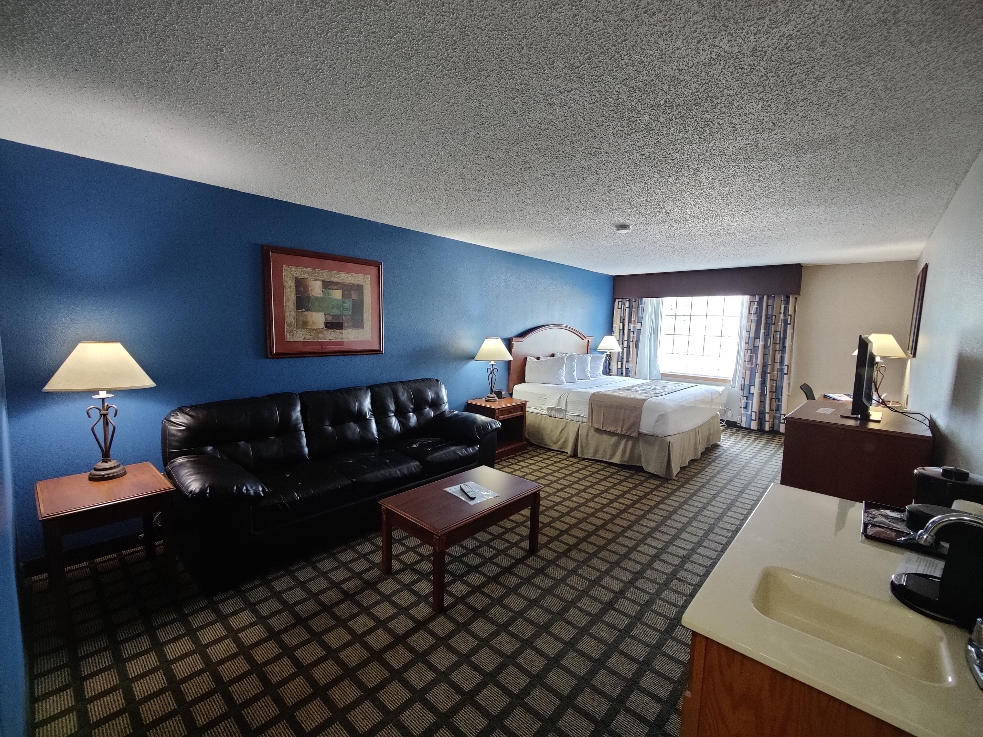 Habitación de hotel con sofá de cuero negro, una mesita de centro de madera, cama con colcha blanca, un pequeño escritorio con lámpara y baño con lavabo y espejo.