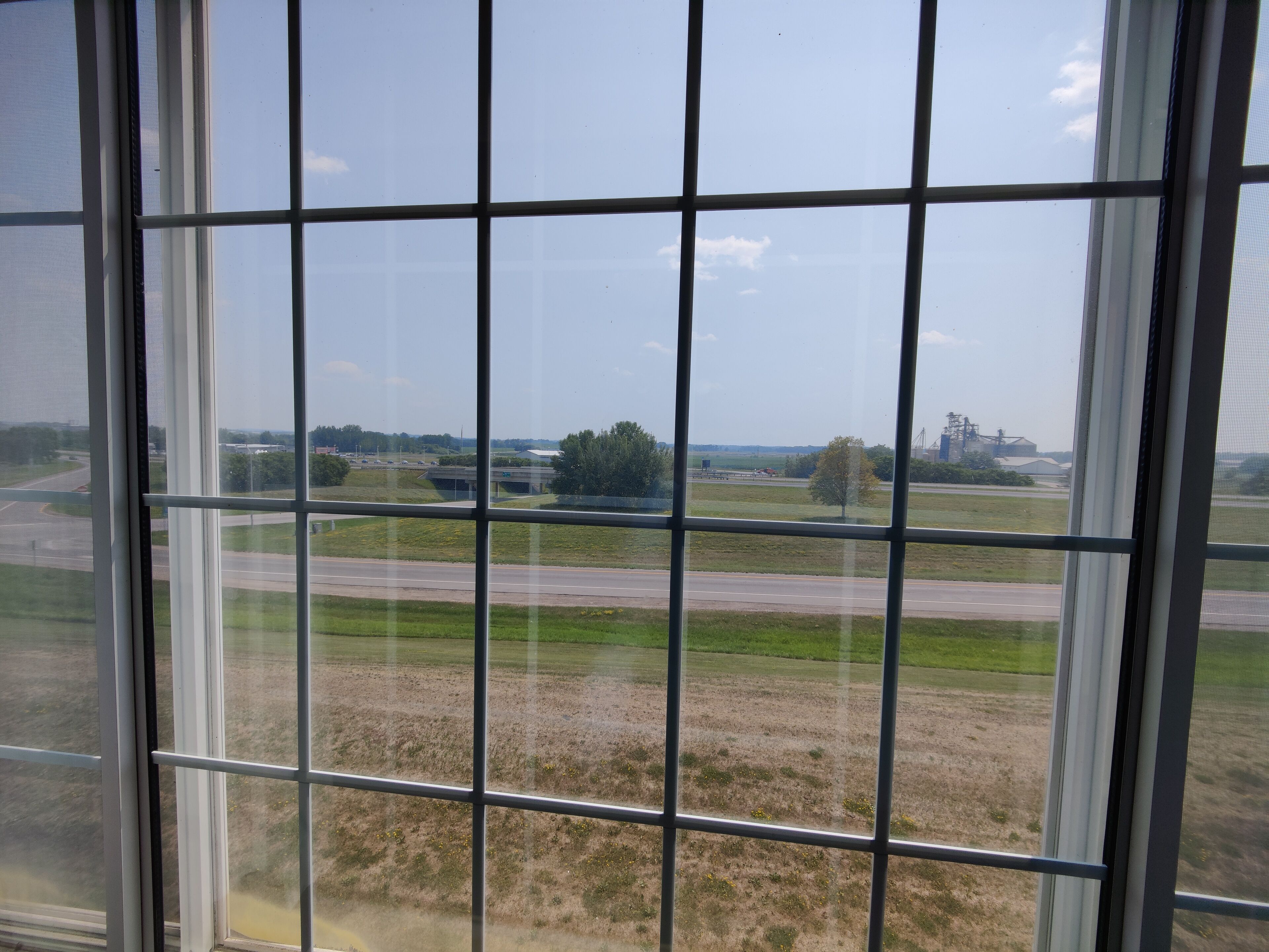 Blick durch ein Fenster mit Gittermuster, das eine Straße, Grünflächen und entfernte Gebäude zeigt.