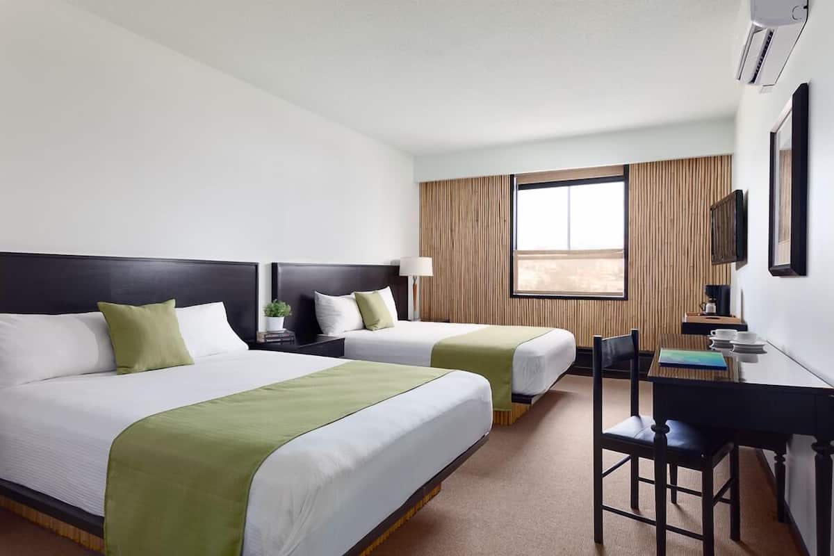 Une chambre d’hôtel avec deux lits, un bureau, une chaise, une lampe et une grande fenêtre avec des rideaux.