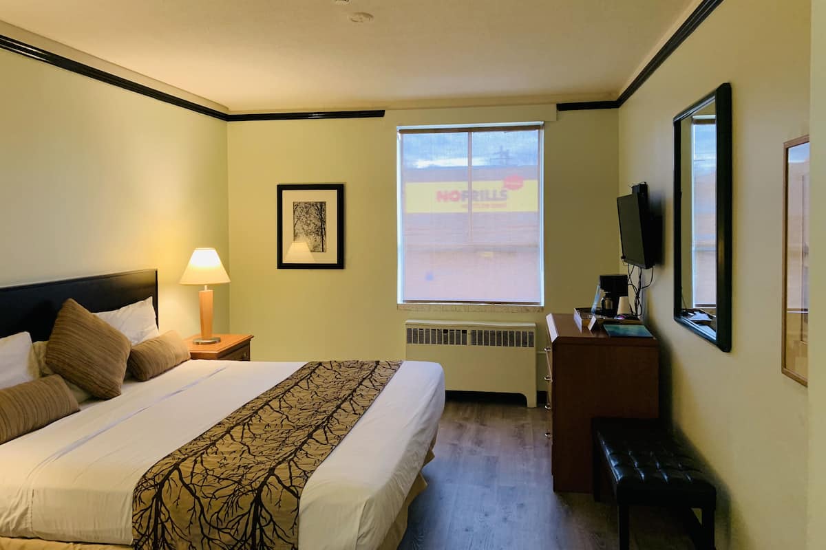 Une chambre d’hôtel avec un lit, une table de chevet, une lampe, un tableau encadré, une fenêtre donnant sur un immeuble et un téléviseur posé sur un meuble.