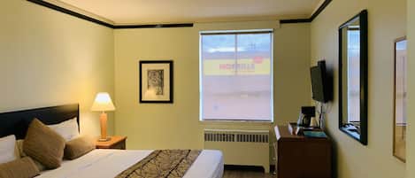 A hotel room with a bed, a nightstand, a lamp, a framed picture, a window with a view of a building, and a television on a stand.