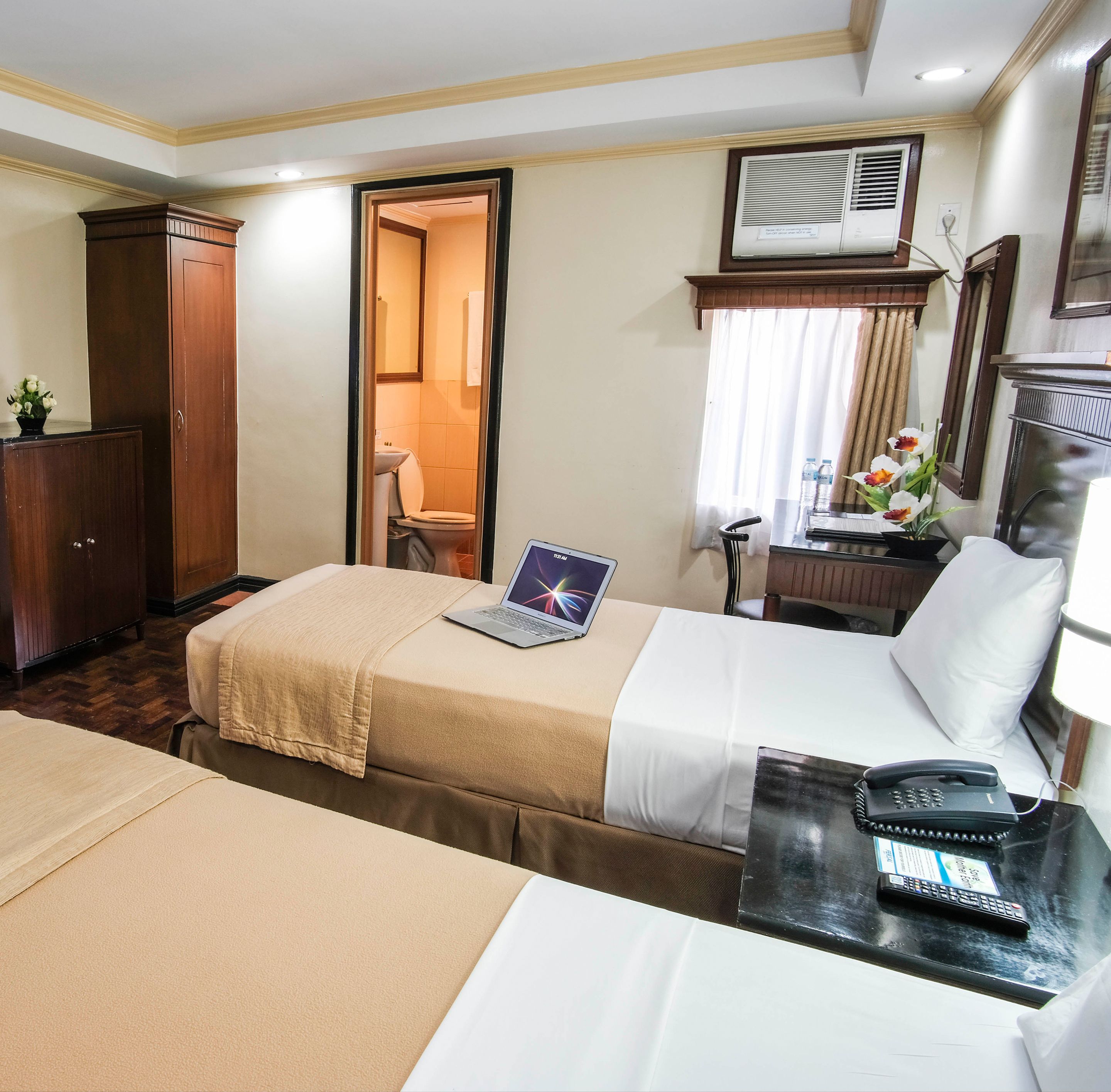 A hotel room with a bed, a desk with a laptop, a telephone, and a bathroom visible through an open door.
