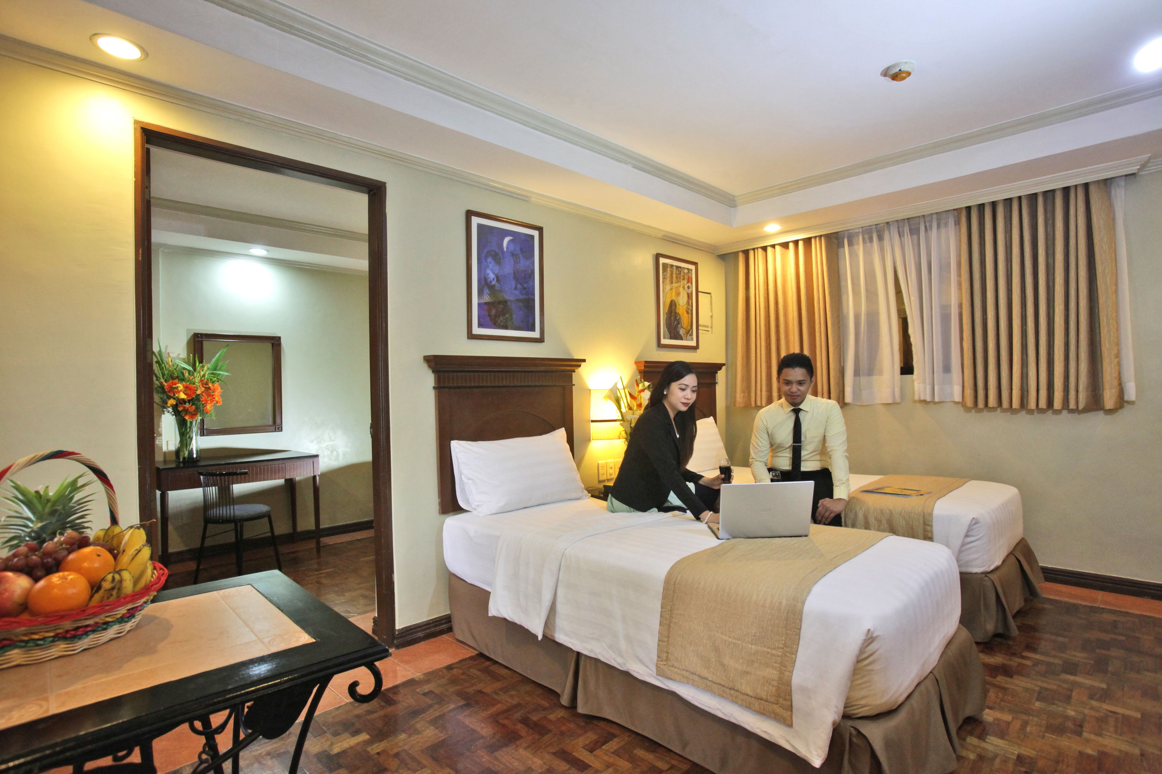 A hotel room with two beds, a dining table with fruit, and a desk with a lamp.