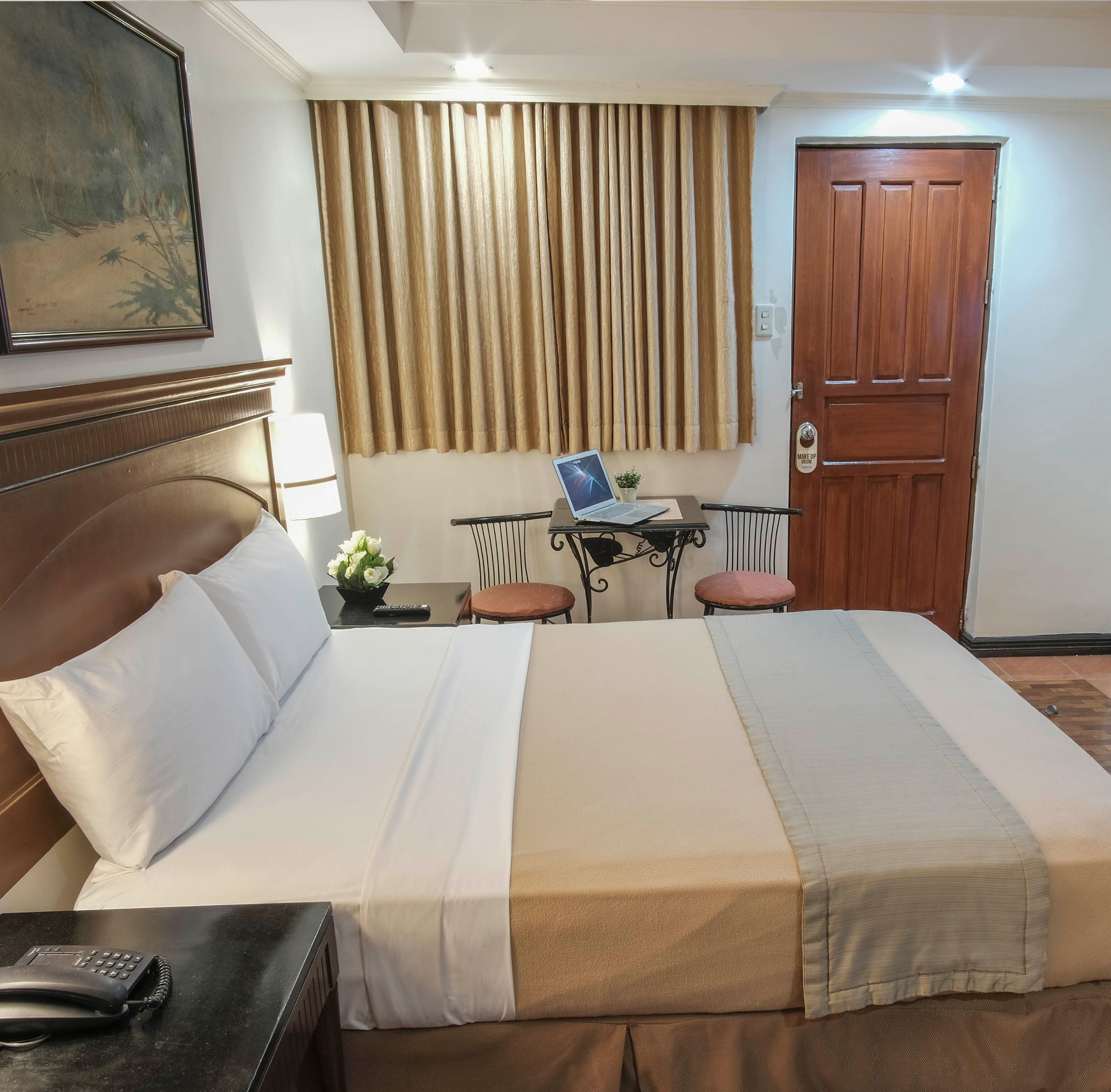A hotel room with a large bed, a desk with a laptop, a chair, a small table with a plant, and a door with a "Make Up Room" sign.
