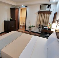 Habitación de hotel con cama, escritorio, silla, armario, baño visible a través de una puerta abierta, una planta en maceta y un espejo.
