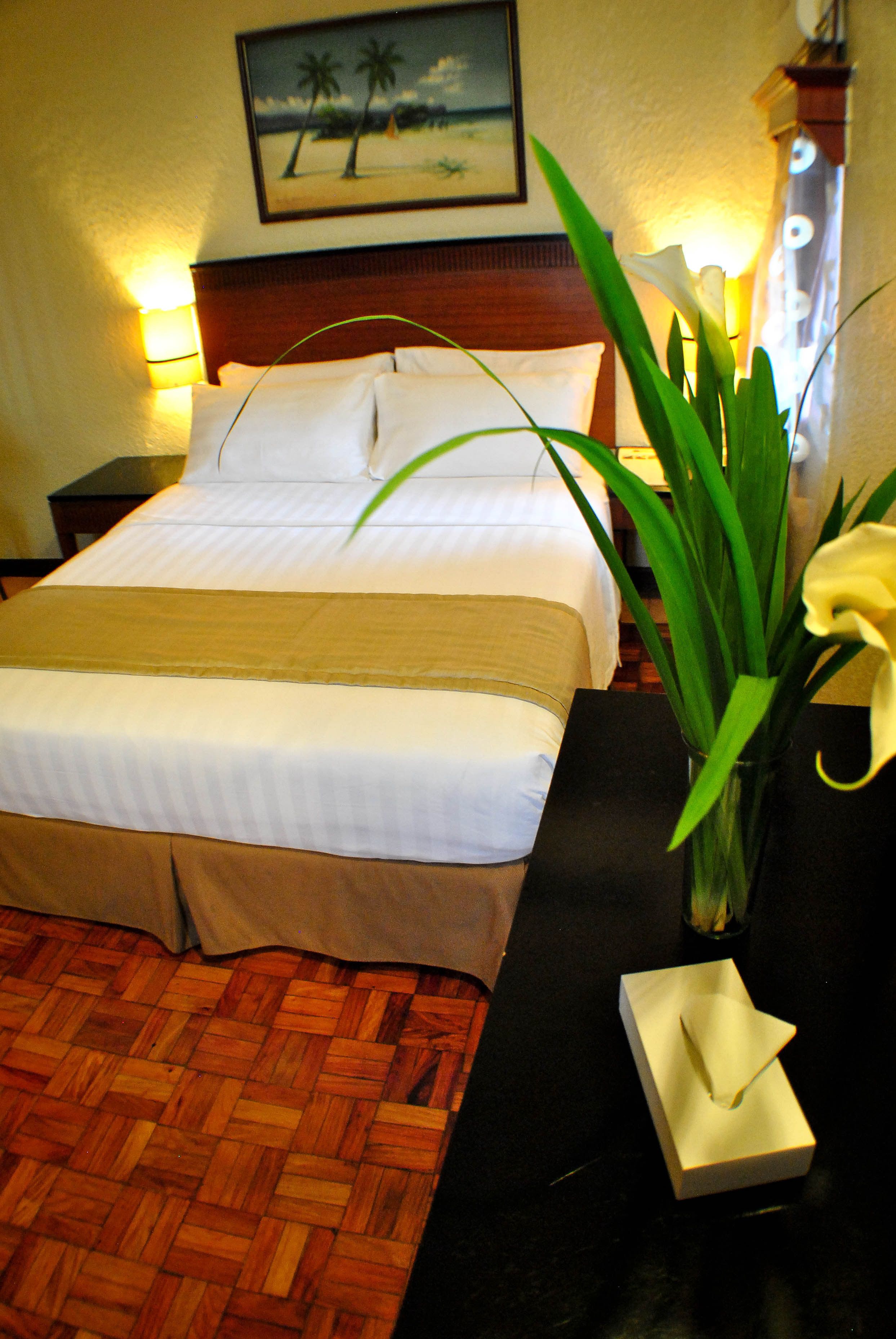 A neatly made bed with a wooden headboard, a nightstand with a lamp, a vase with flowers, and a painting on the wall.
