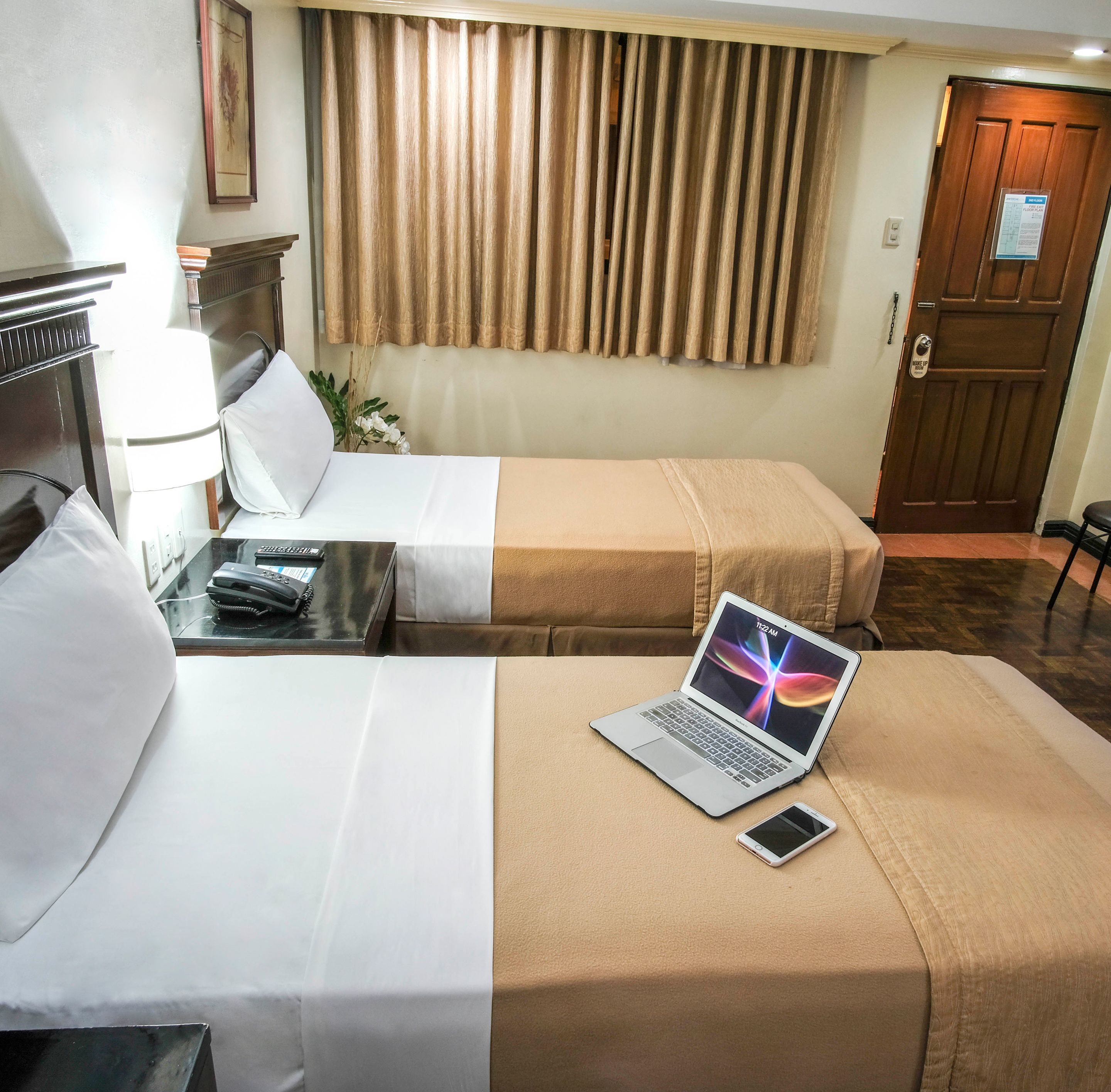 Habitación de hotel con dos camas, un escritorio con una computadora portátil y un teléfono, una lámpara y una puerta con un letrero.