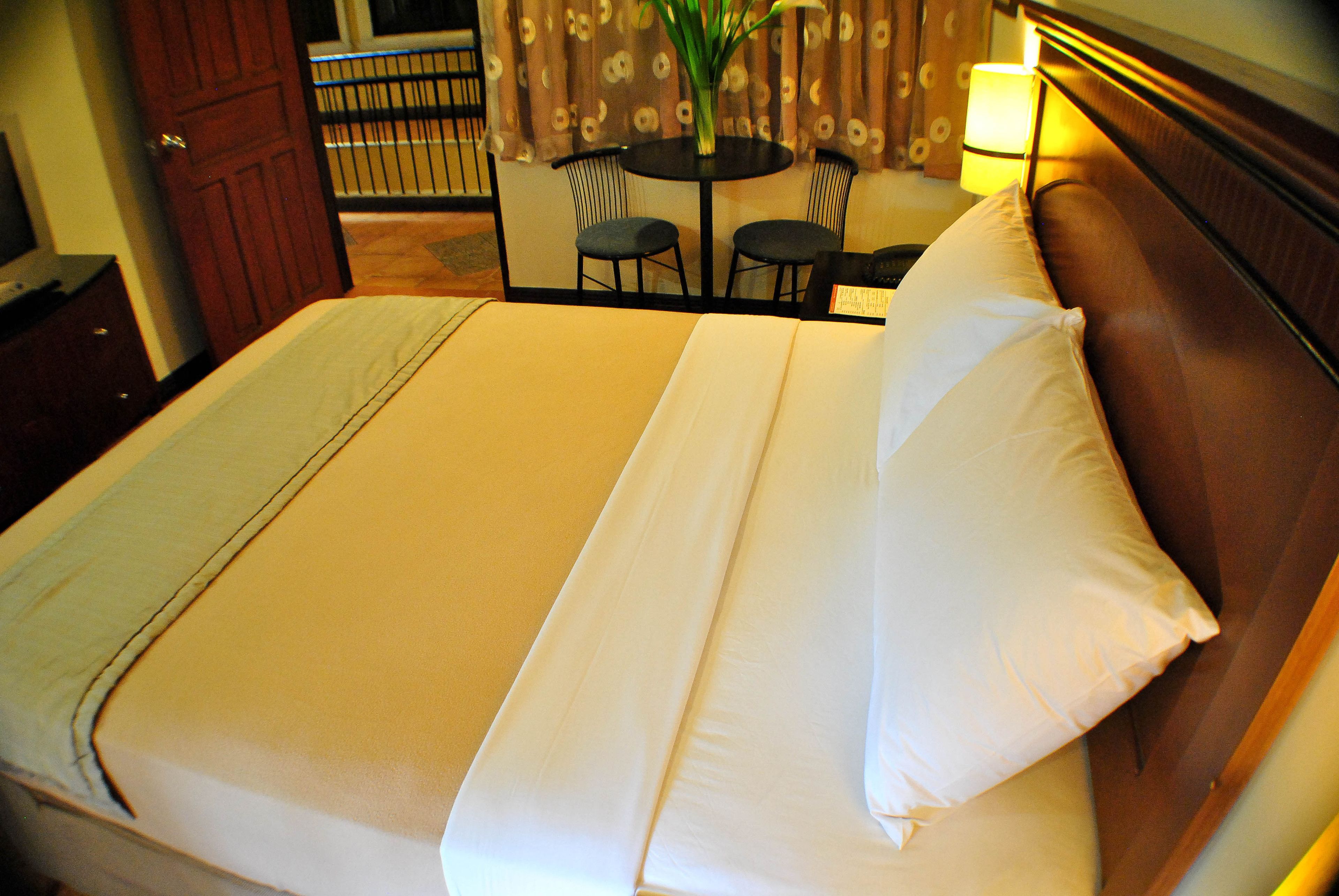 A neatly made bed with a white comforter and pillows, a wooden headboard, and a small table with chairs in the background.
