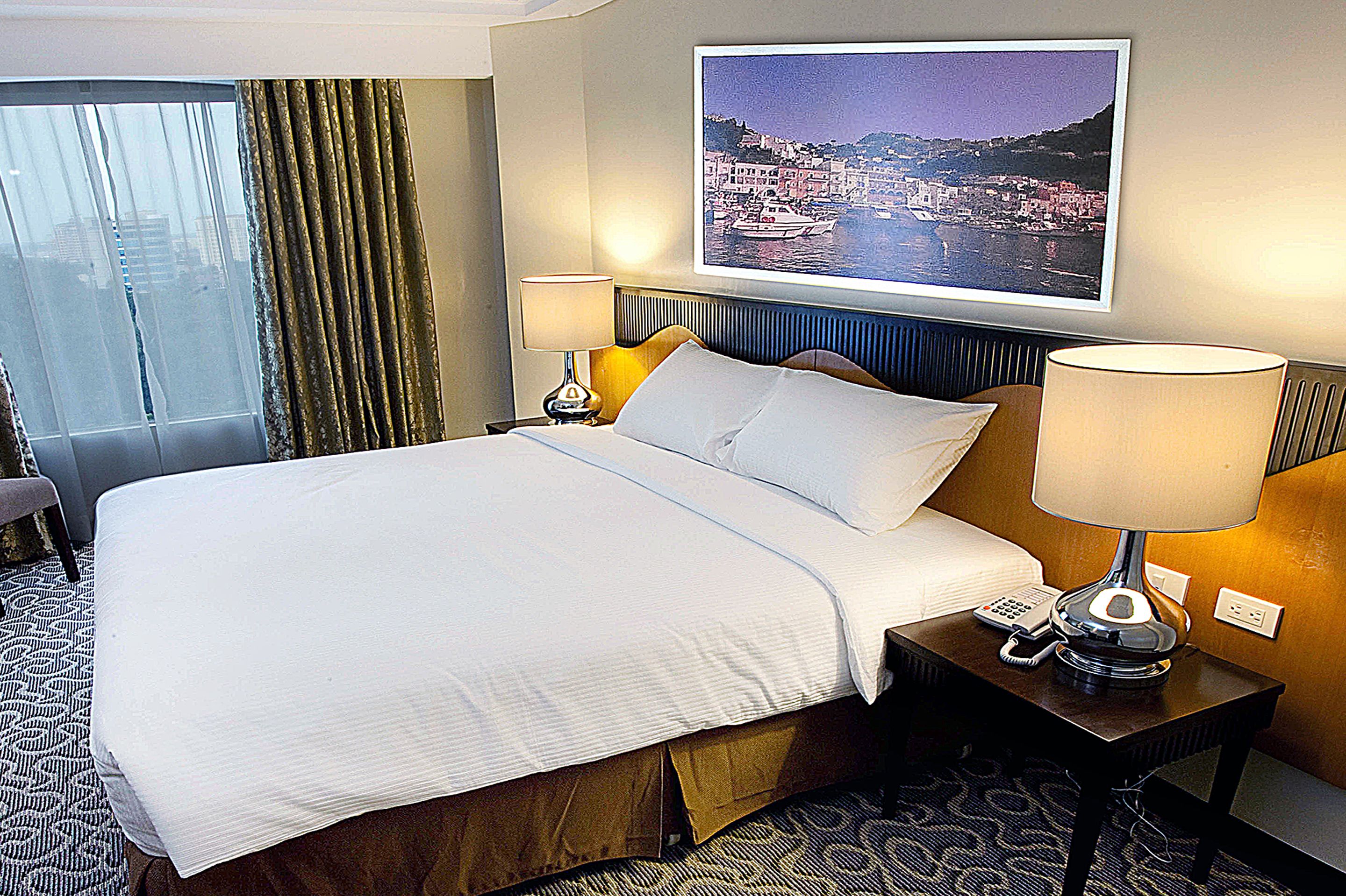 A hotel room with a large bed, two bedside tables with lamps, a window with curtains, and a cityscape painting on the wall.