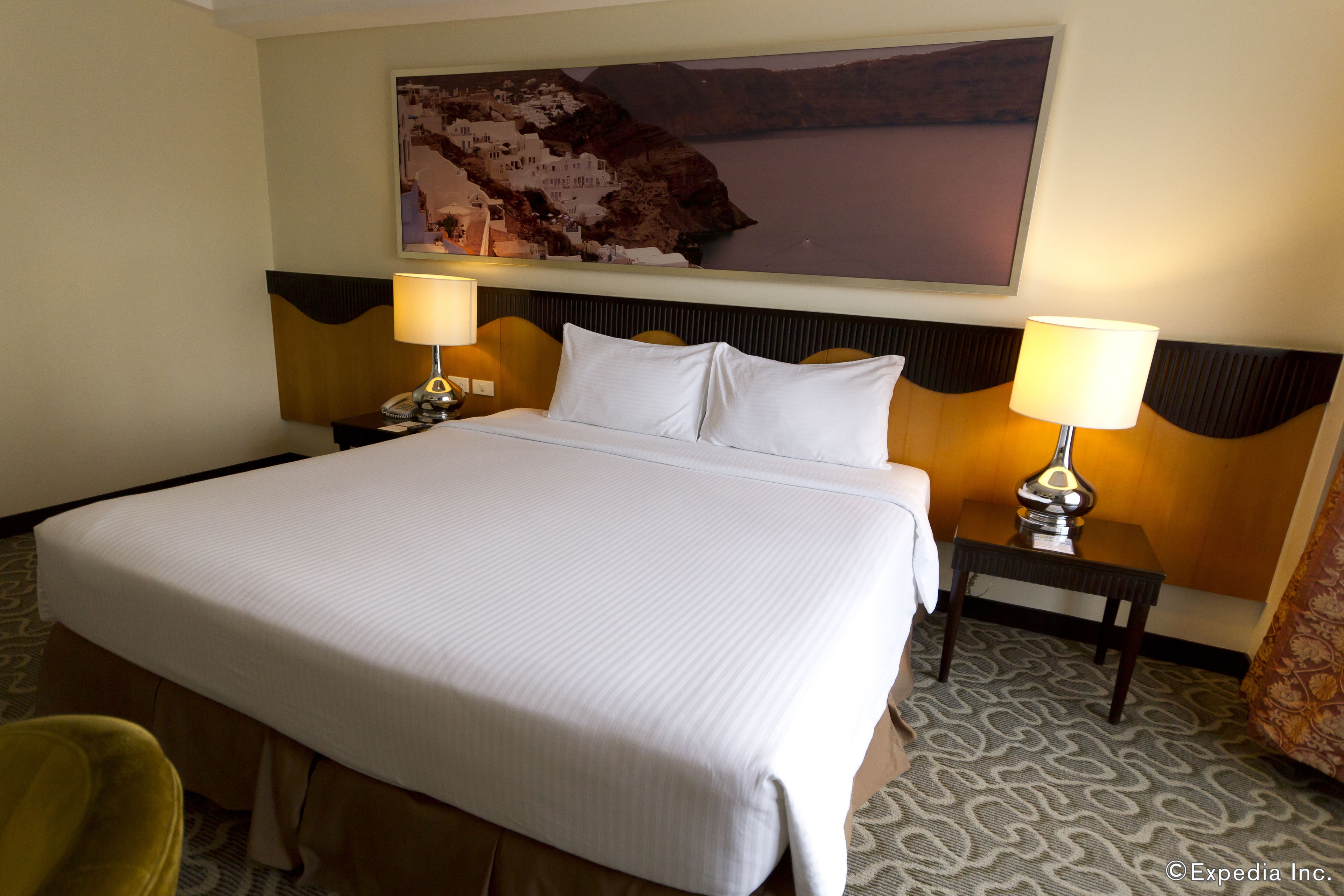 A hotel room with a large bed, two bedside lamps, a headboard with a unique design, a wall-mounted painting, and a patterned carpet.