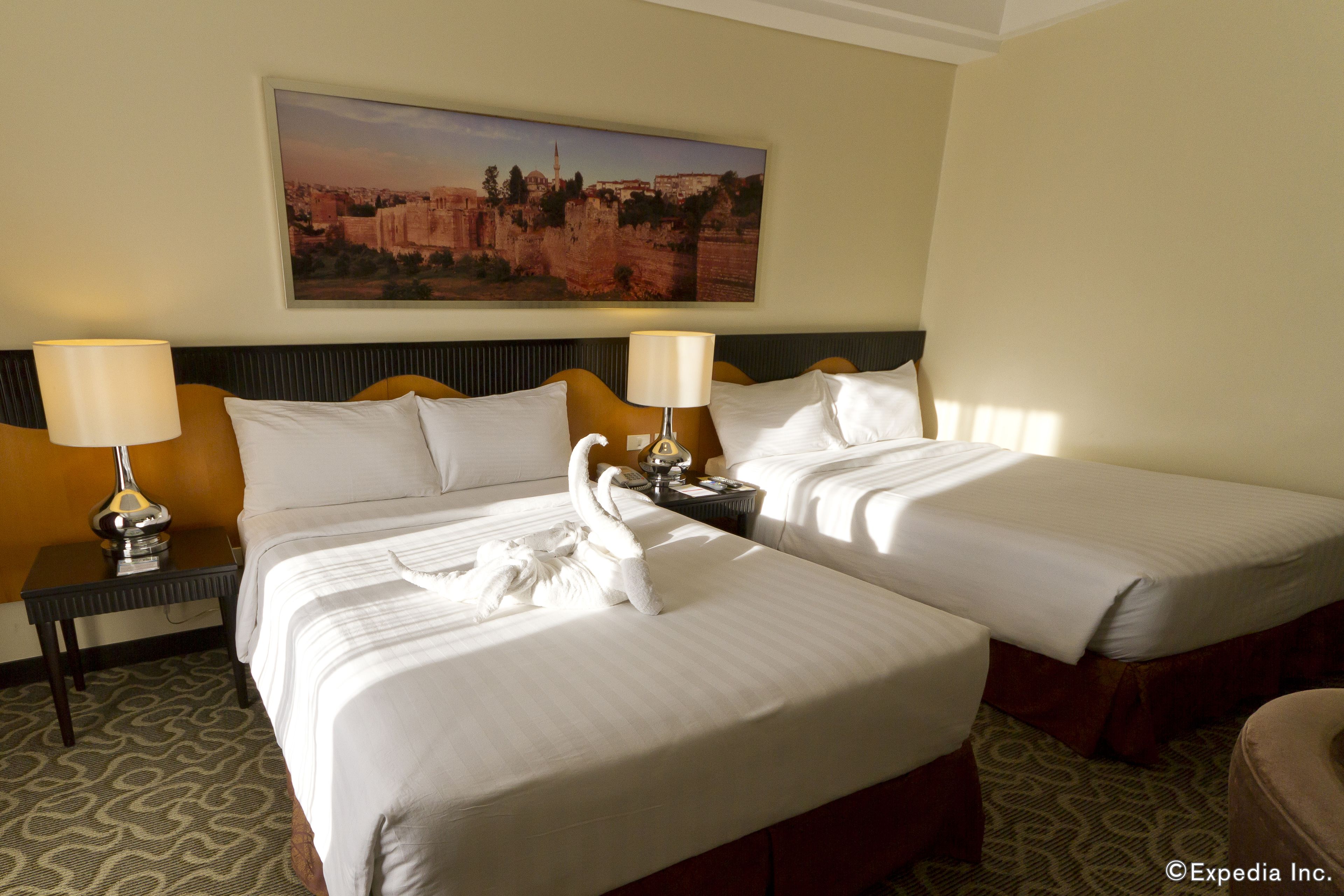 A hotel room with two beds, a nightstand with lamps, a painting on the wall, and a patterned carpet.