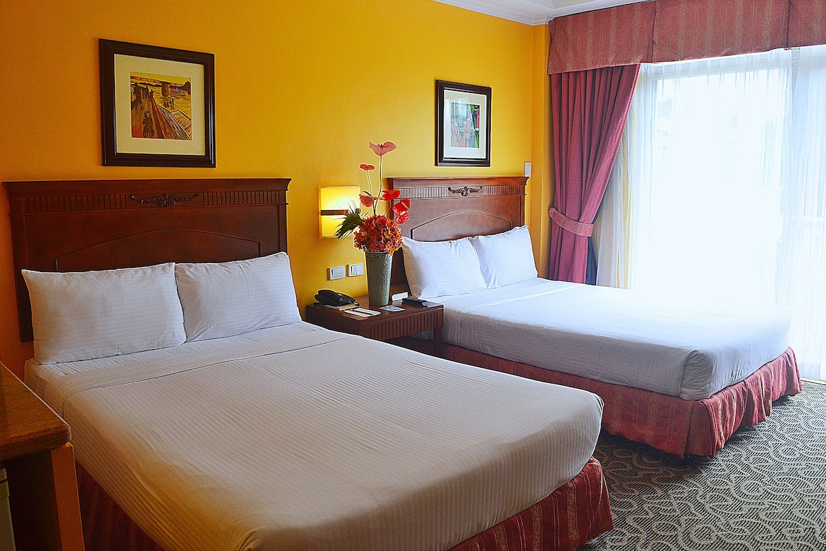 A hotel room with two beds, a nightstand, a vase with flowers, and two framed pictures on the wall.