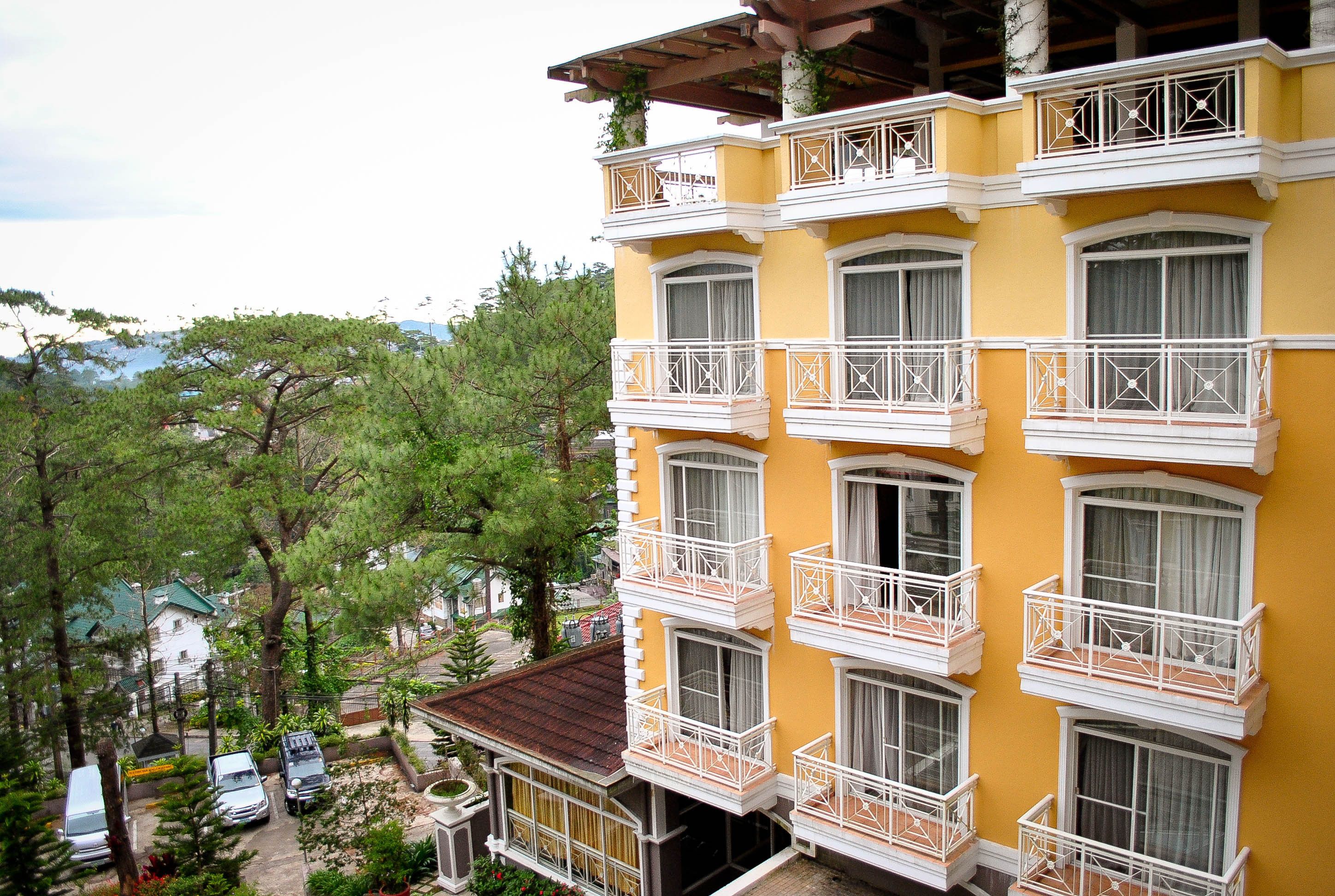 A multi-story building with balconies and large windows, surrounded by trees and parked cars.