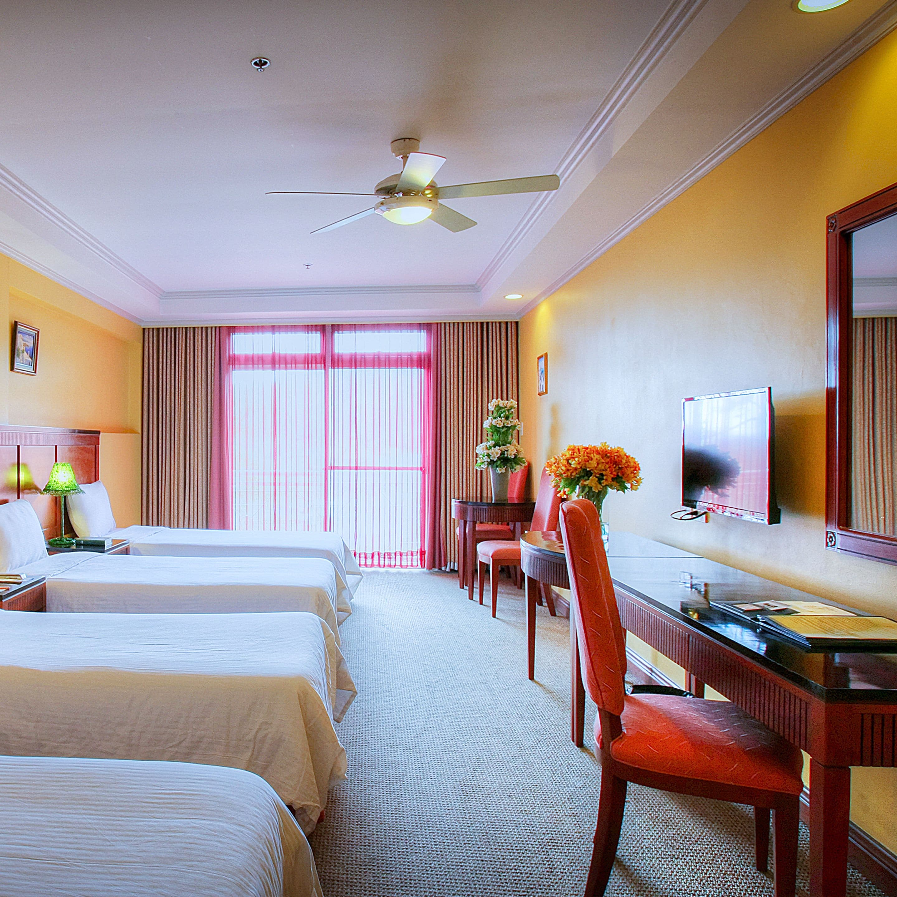 A hotel room with two beds, a desk with a chair, a television, and a ceiling fan.