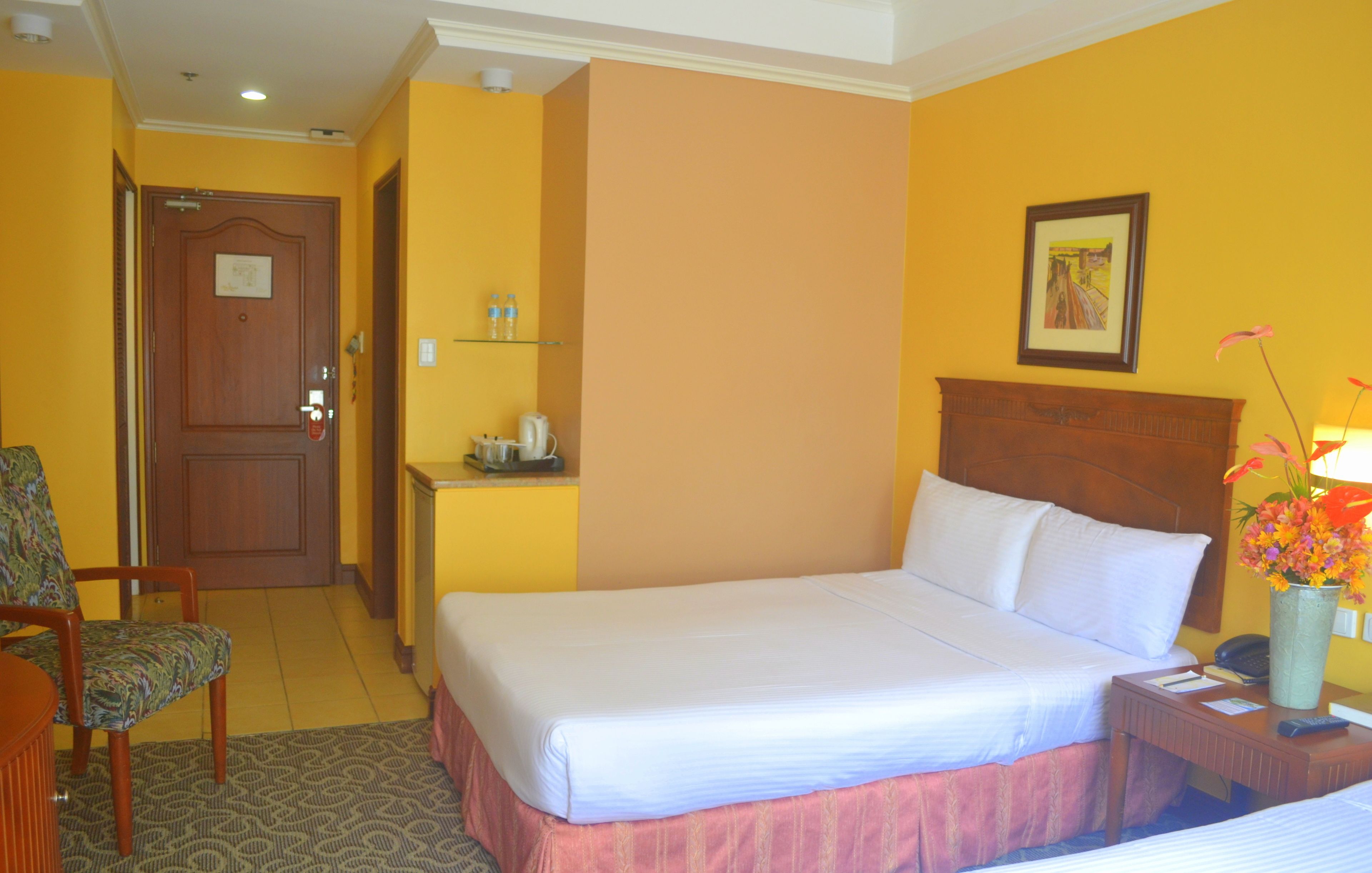 A hotel room with a bed, a chair, a desk with a lamp, and a vase with flowers.