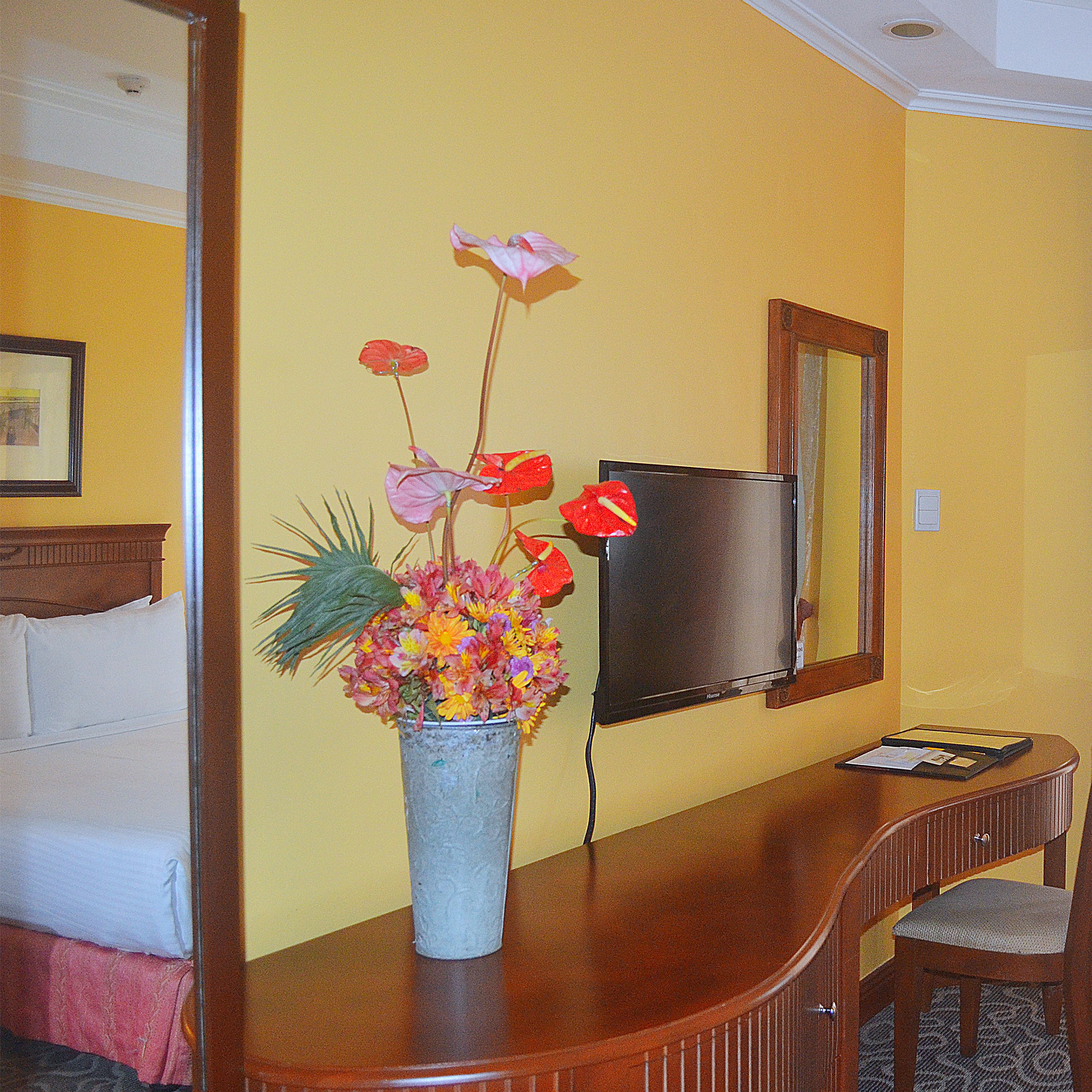 Une chambre d’hôtel avec un bureau en bois, un vase rempli de fleurs, une télévision et un lit avec des oreillers.