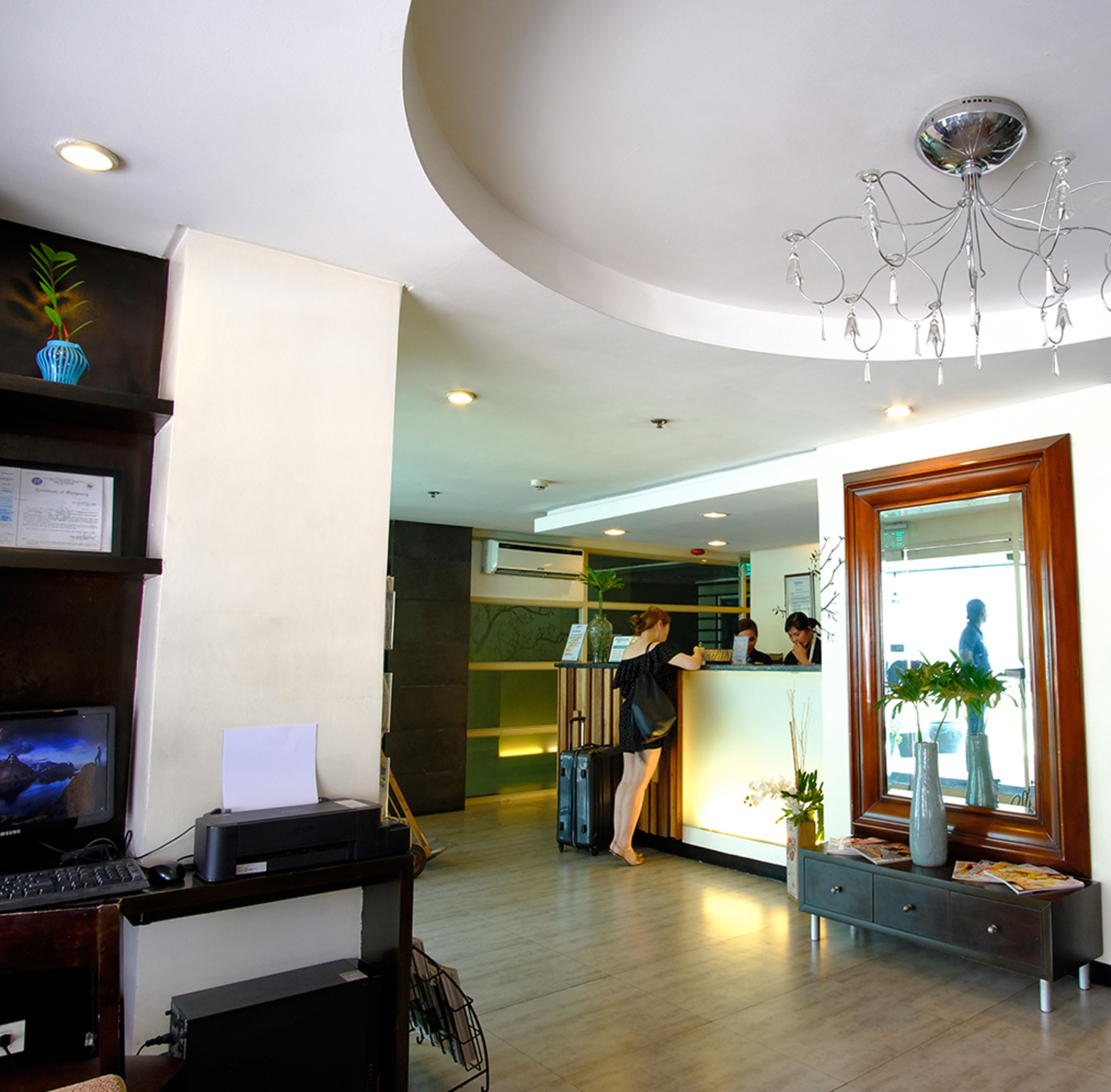A reception area with a desk, a computer, and a large mirror. There are plants and a chandelier.