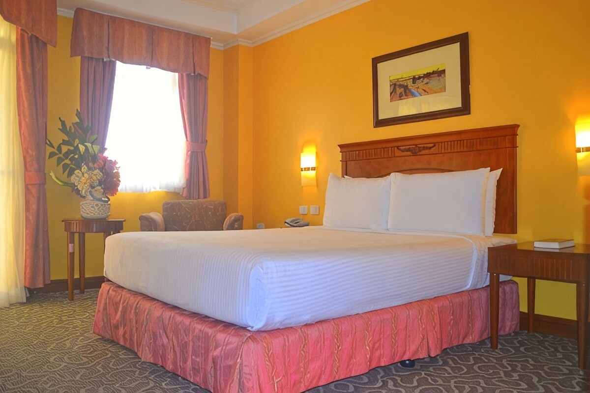 A hotel room with a large bed, a chair, a small table, a vase with flowers, and a painting on the wall.