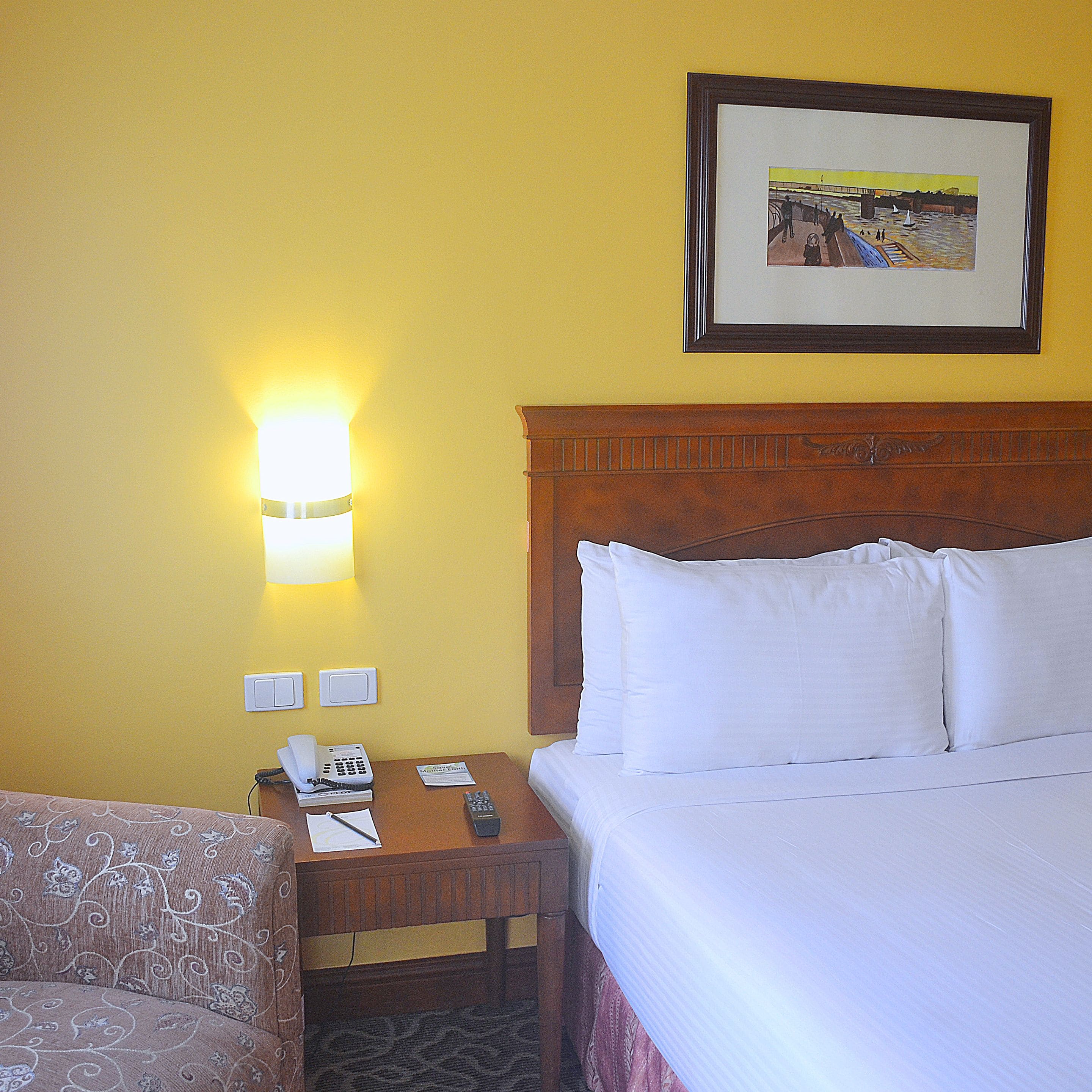 A hotel room with a bed, a nightstand with a phone and remote, a lamp, and a framed picture on the wall.