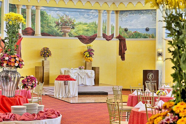 Un salón de banquetes con mesas dispuestas para un evento formal, un escenario y un mural en la pared.