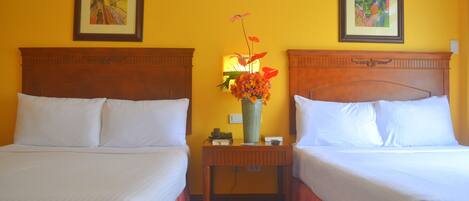 Two double beds with white linens, a wooden nightstand with a vase of flowers, and two framed pictures on the yellow walls.