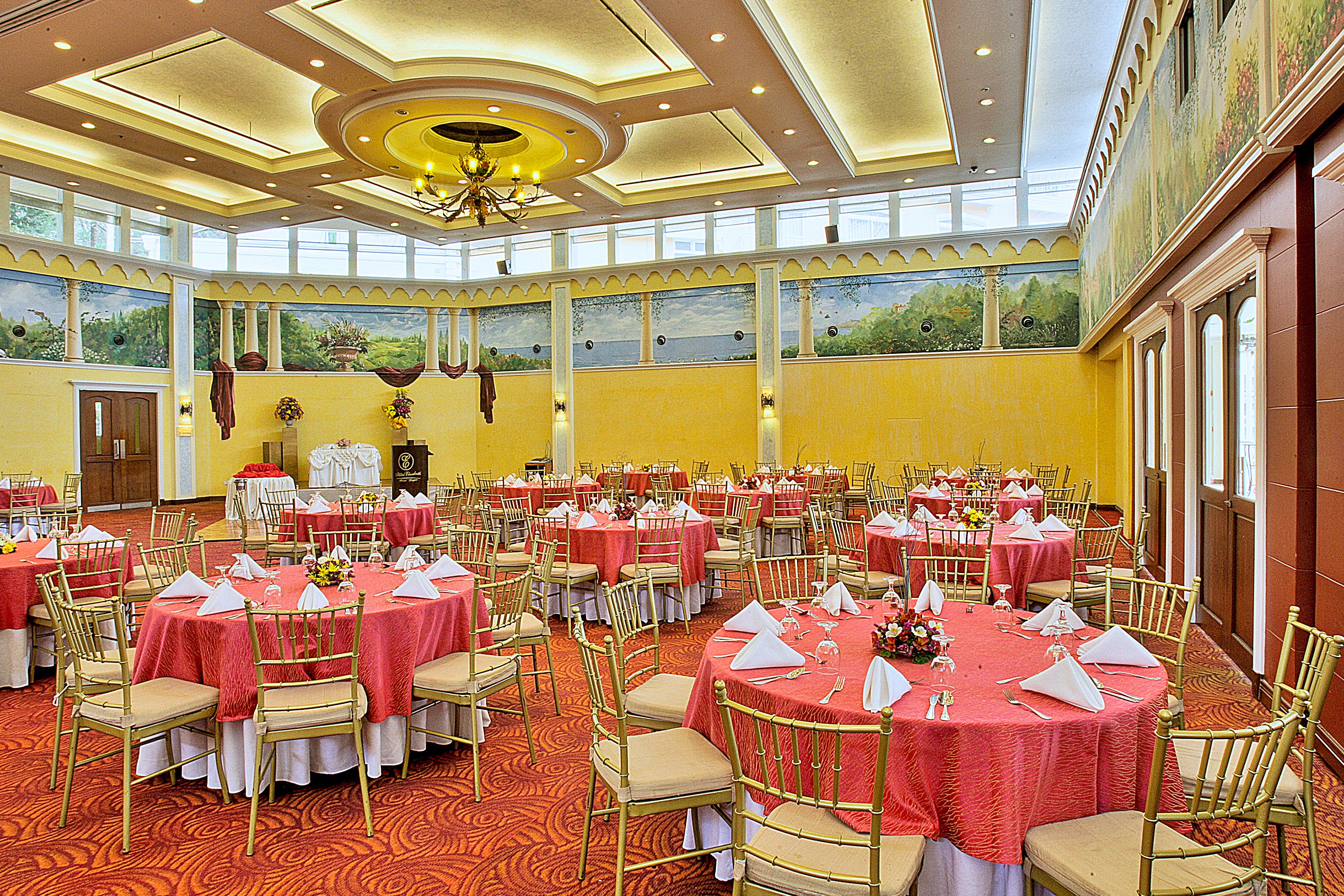 Une salle de banquet dotée de nombreuses tables rondes, chacune recouverte d’une nappe blanche, agrémentée de serviettes rouges et entourée de chaises dorées.