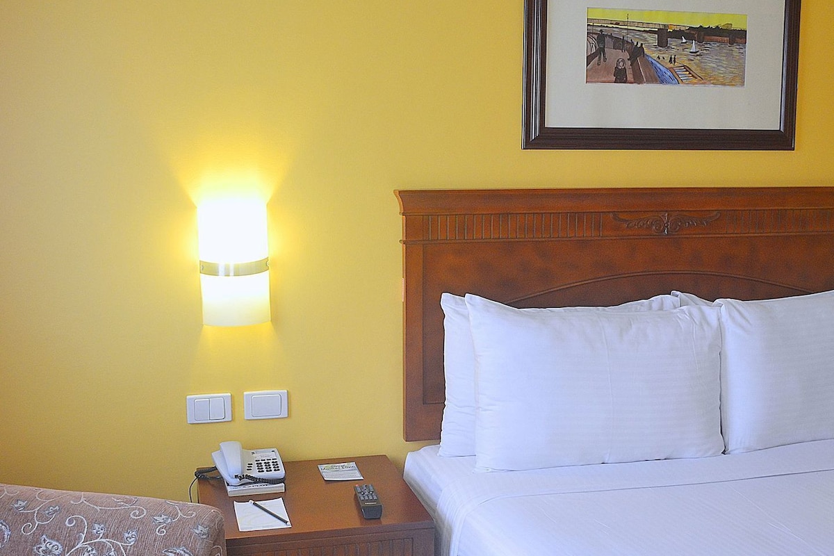 A hotel room with a bed, a nightstand with a phone and remote, a lamp, and a framed picture on the wall.