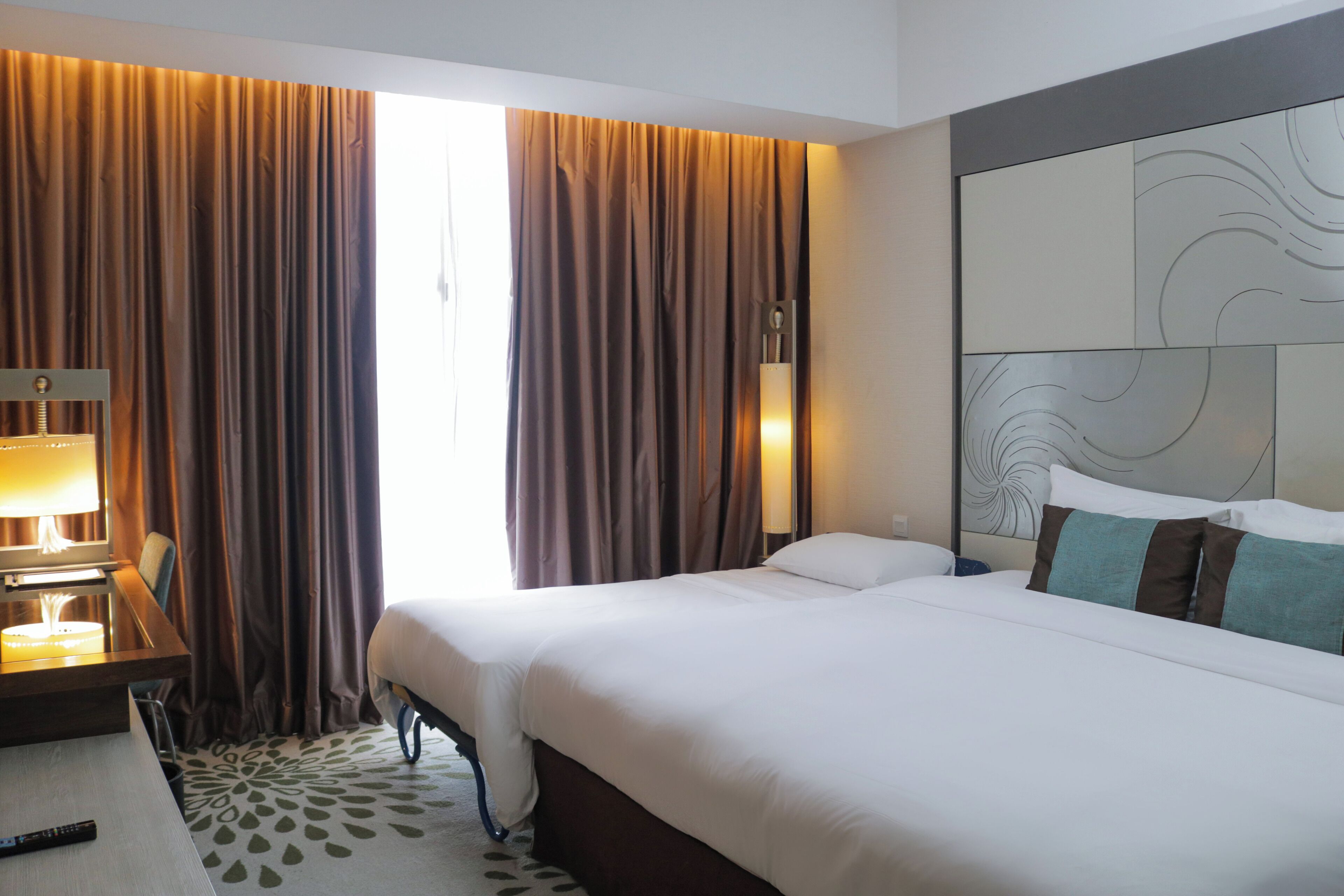 A hotel room with a bed, a desk with a lamp, curtains, and a headboard with a decorative panel.