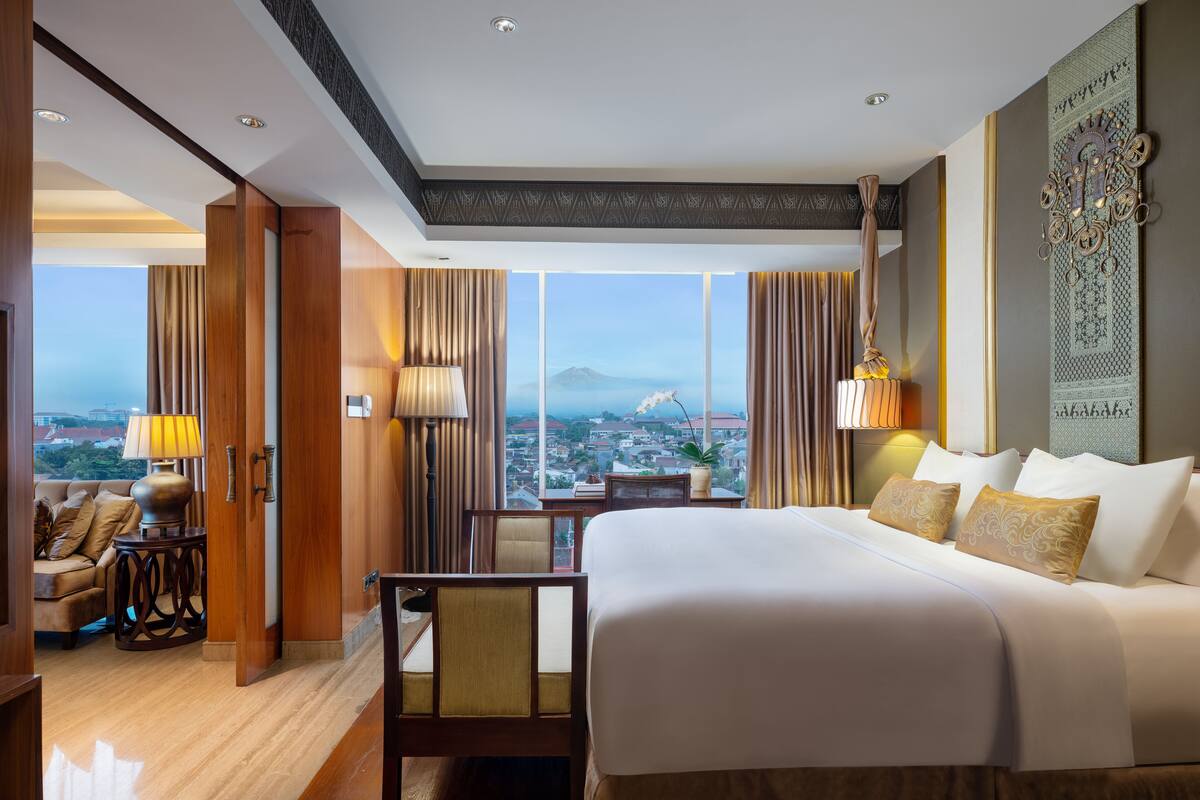 Habitación de hotel con una cama grande, un área de descanso con una silla y una mesa, y vistas de un paisaje urbano a través de amplias ventanas.