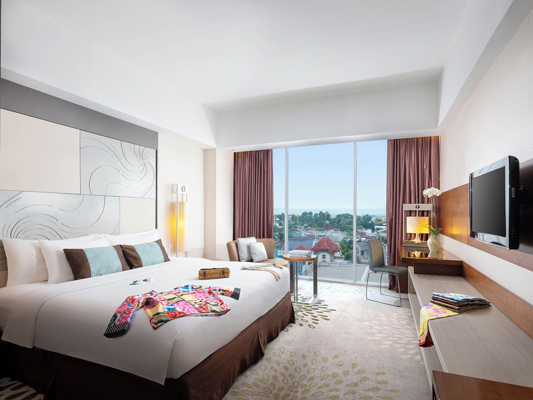 A hotel room with a large bed, a desk, a chair, a TV, and a view of the city.