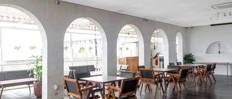 A spacious indoor area with wooden tables and chairs, large windows, and potted plants.