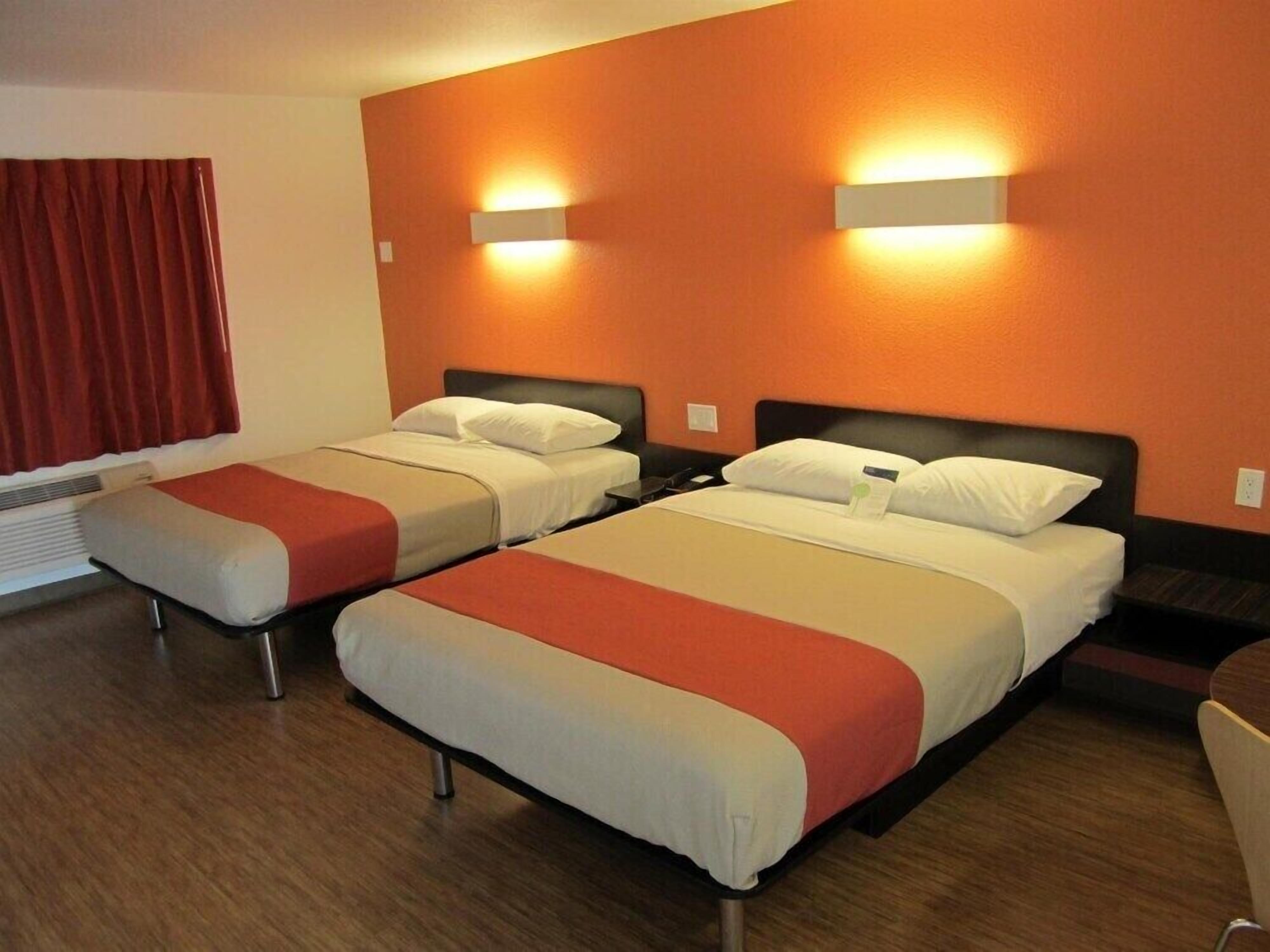 A hotel room with two beds, each with a headboard and bedside tables. There is a window with red curtains and a wall-mounted light fixture.