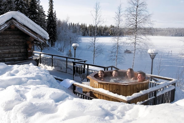 A wooden hot tub surrounded by snow, with a wooden cabin and a snowy landscape in the background.