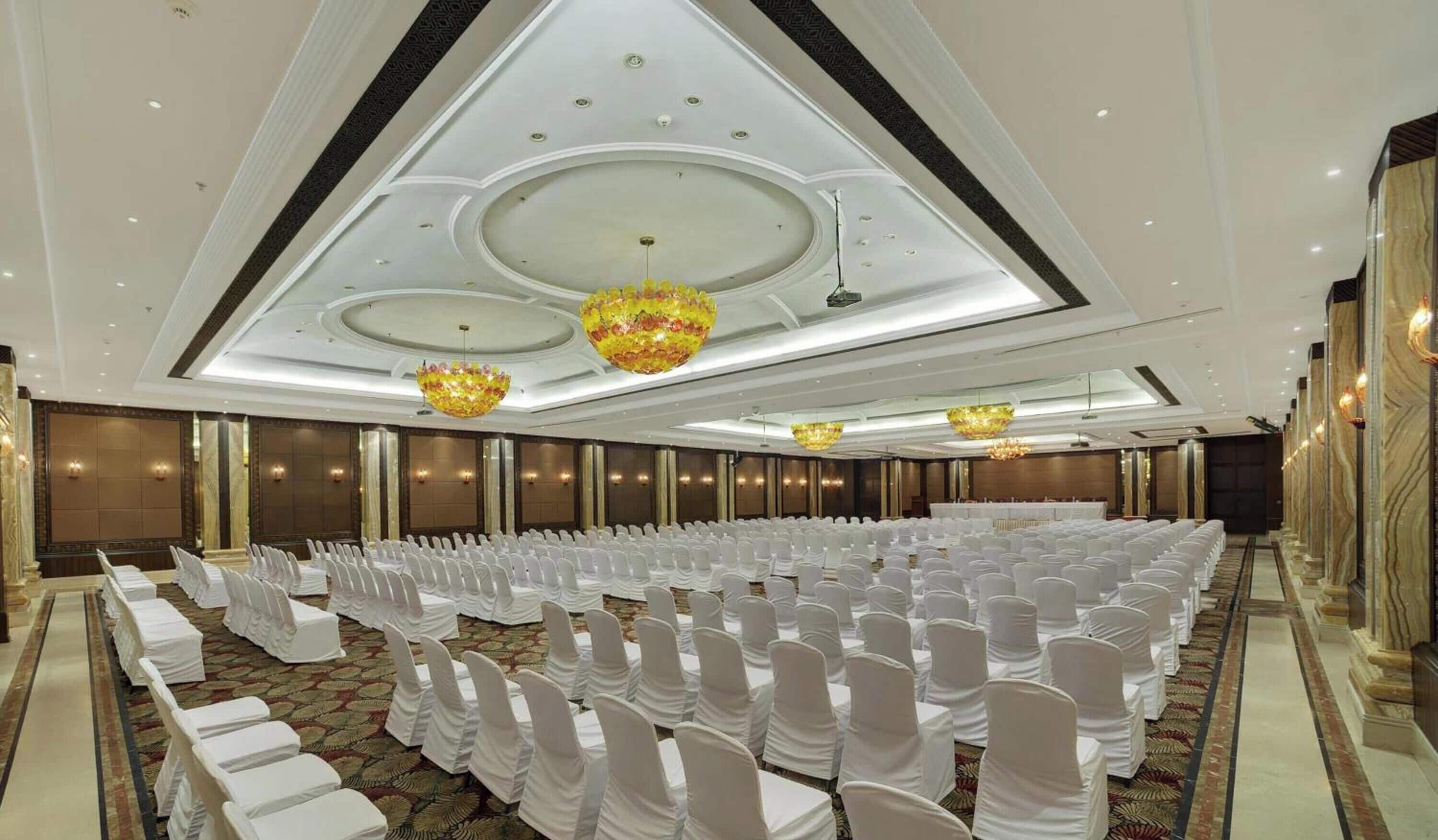 A large conference room with rows of chairs covered in white chair covers, a high ceiling with recessed lighting, and decorative chandeliers.