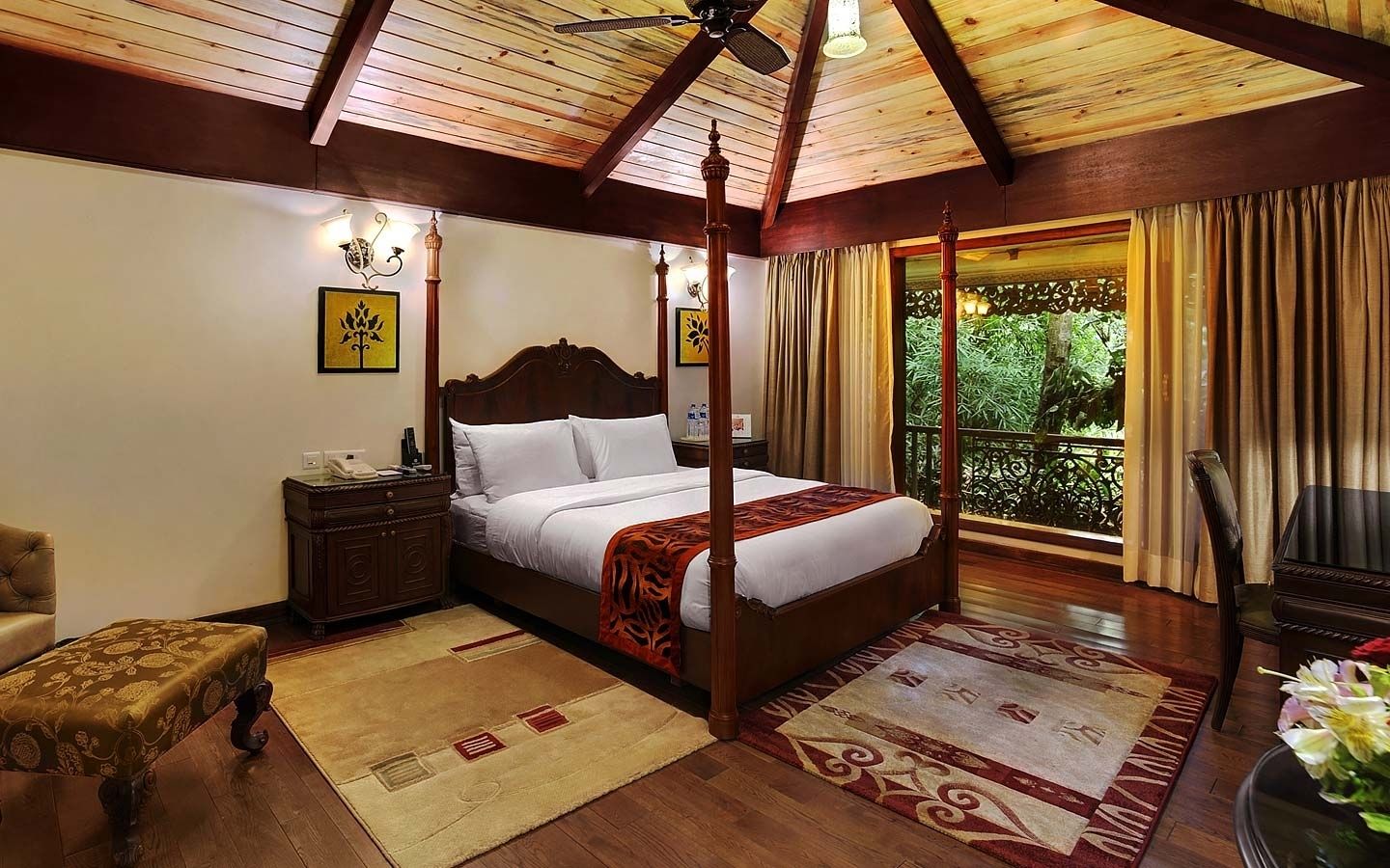 A four-poster bed with white linens, a wooden nightstand with a lamp, a wooden chair, and a view of greenery through a balcony door.