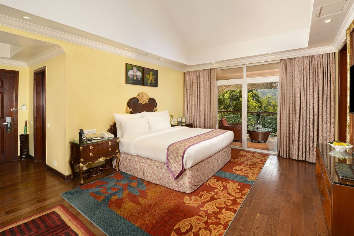 A hotel room with a large bed, a television, a desk with a computer, and a balcony with a view of greenery.
