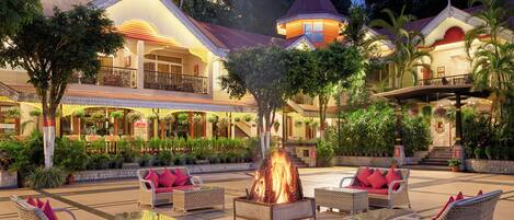 A hotel courtyard with wicker furniture, a central fire feature, and a well-lit building with balconies and potted plants.