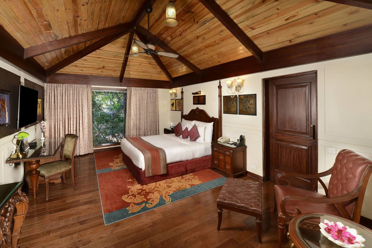 A bedroom with a wooden ceiling, a bed with a red bedspread, a desk with a lamp, and a chair.
