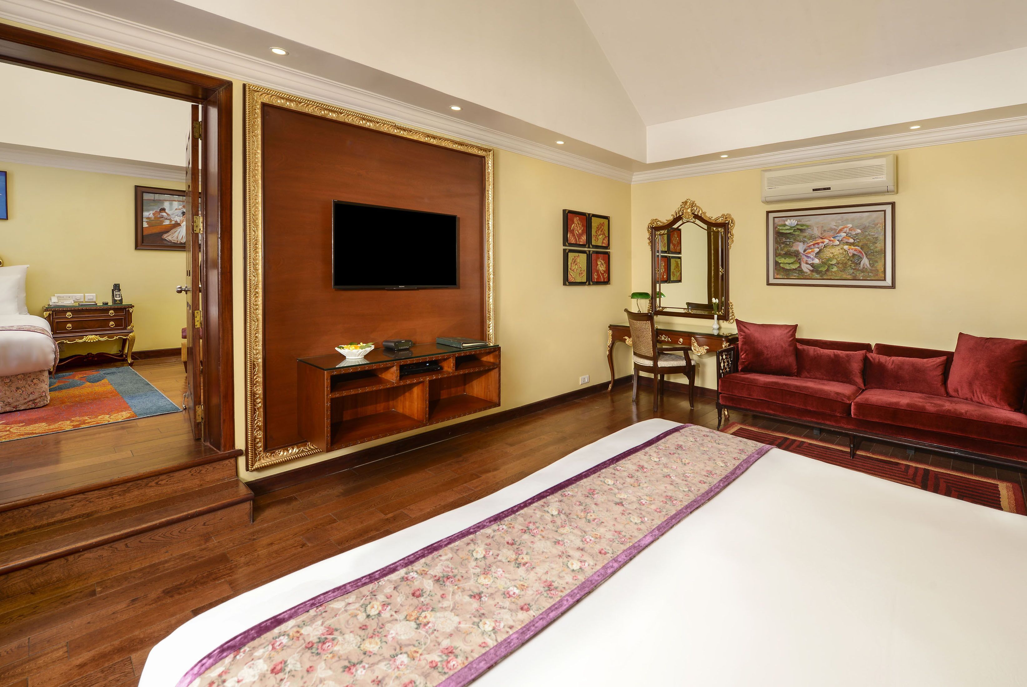A hotel room with a large bed, a TV mounted on a wooden panel, a red sofa, a mirror, and a wooden cabinet.