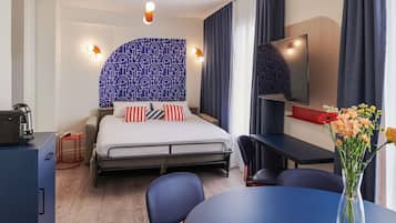 A hotel room with a bed, bedside table, desk, chair, and a vase with flowers.