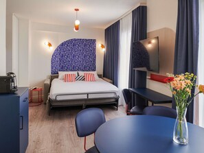 Quarto de hotel com cama, mesa de cabeceira, escrivaninha, cadeira e um vaso com flores.