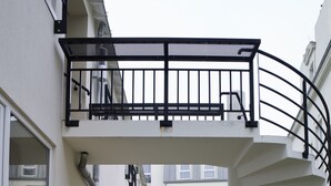 Un sendero de madera conduce a una escalera en espiral con barandilla negra, rodeado de edificios modernos con grandes ventanales y un área ajardinada.