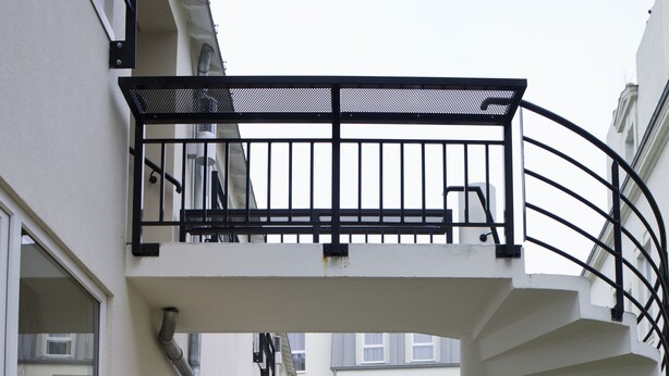 Un sendero de madera conduce a una escalera en espiral con barandilla negra, rodeado de edificios modernos con grandes ventanales y un área ajardinada.