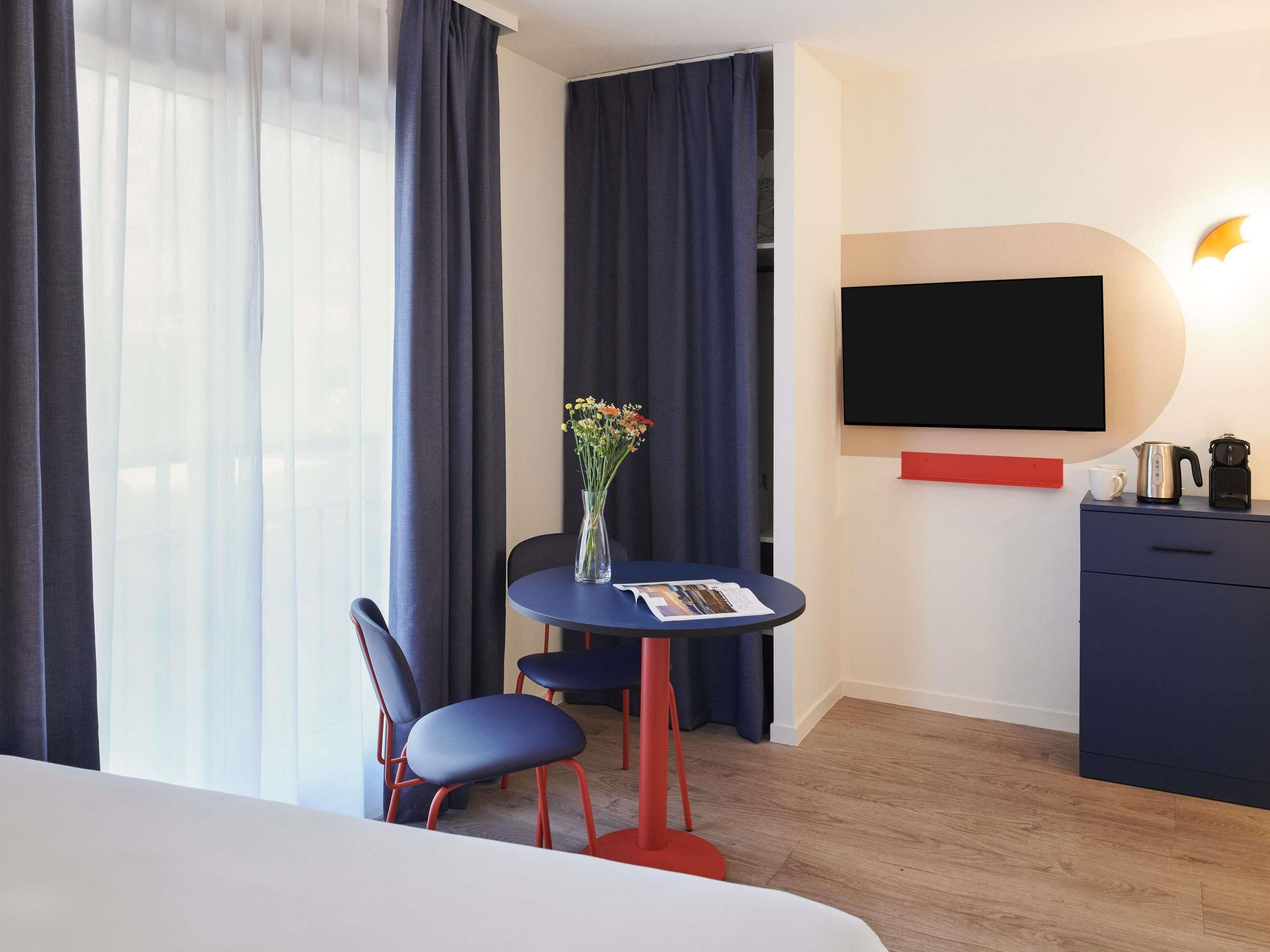 Habitació d'hotel amb llit, escriptori amb cadira, gerro amb flors, televisió de pantalla plana, cafetera i prestatge vermell.