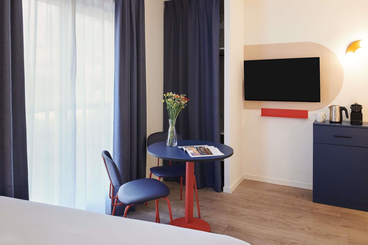 Ein Hotelzimmer mit einem Bett, einem Schreibtisch mit Stuhl, einer Blumenvase, einem Flachbildfernseher, einer Kaffeemaschine und einem roten Regal.