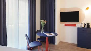 Habitació d'hotel amb llit, escriptori amb cadira, gerro amb flors, televisió de pantalla plana, cafetera i prestatge vermell.