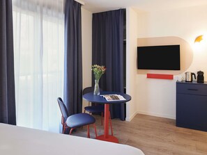 Ein Hotelzimmer mit einem Bett, einem Schreibtisch mit Stuhl, einer Blumenvase, einem Flachbildfernseher, einer Kaffeemaschine und einem roten Regal.